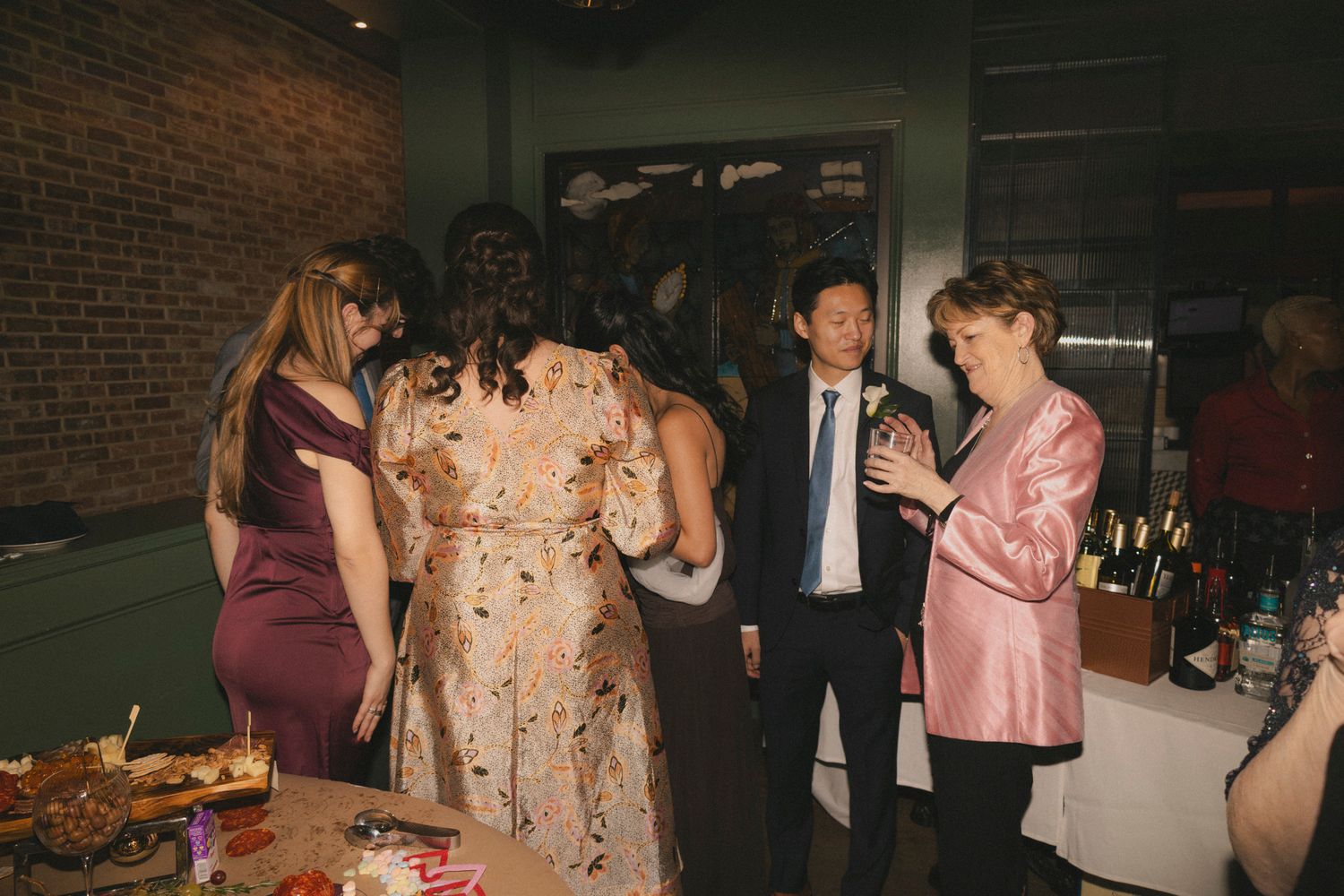 Guests mingle around a food table at an indoor evening event, dressed in formal and semi-formal attire.