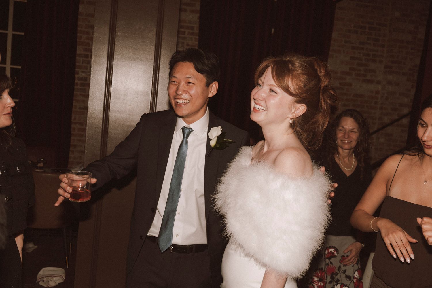Smiling bride in fur stole and groom in teal tie celebrating at lively wedding reception.