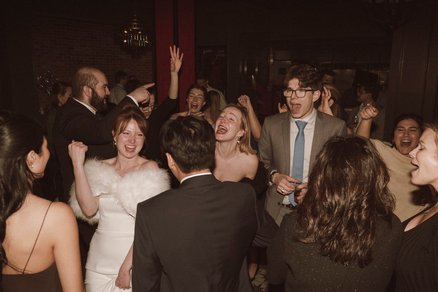 Bride in white gown celebrates joyfully on crowded dance floor with guests at wedding reception.
