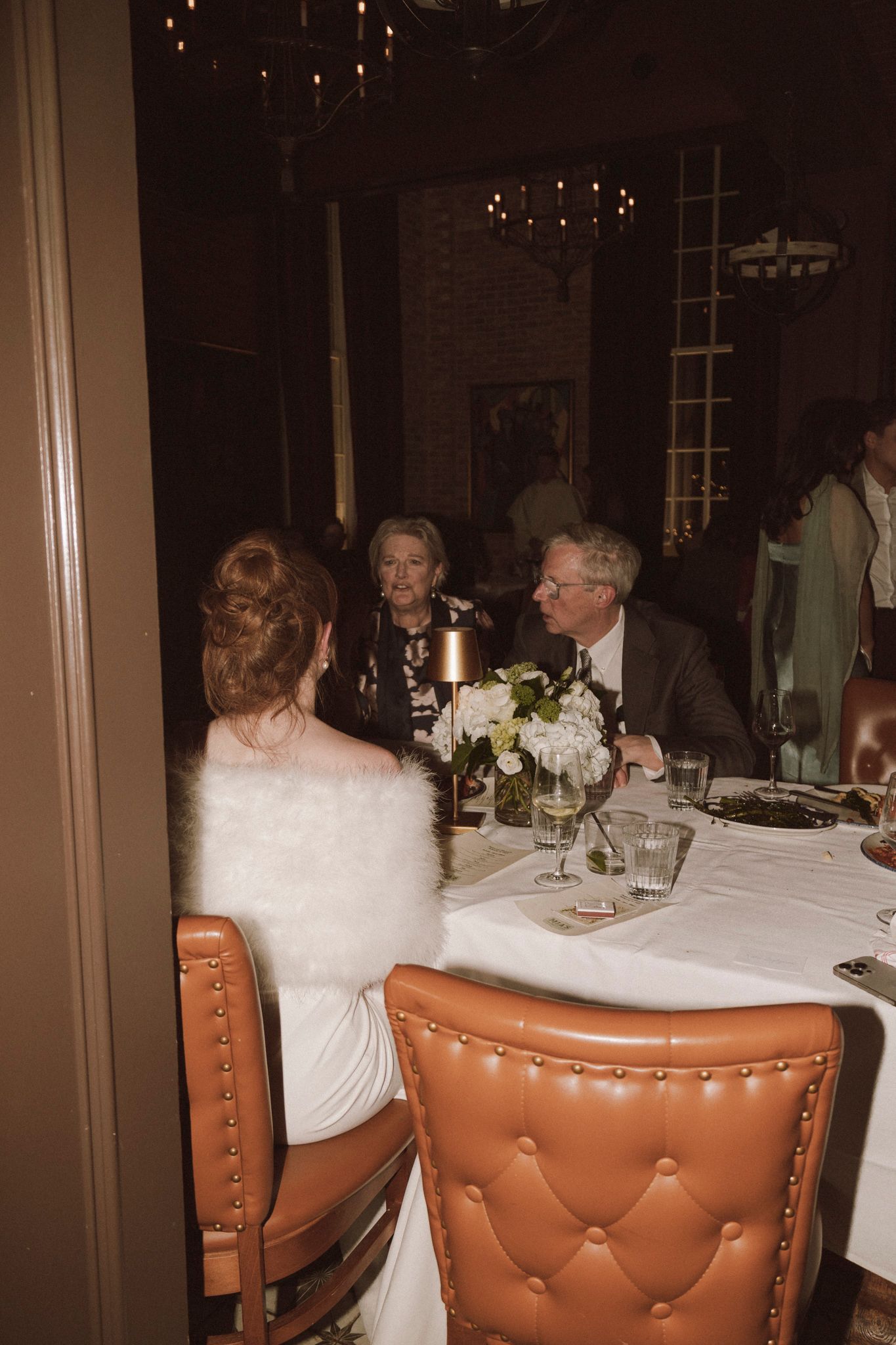 Bride in white dress with feather wrap seated at wedding reception table with older couple guests.