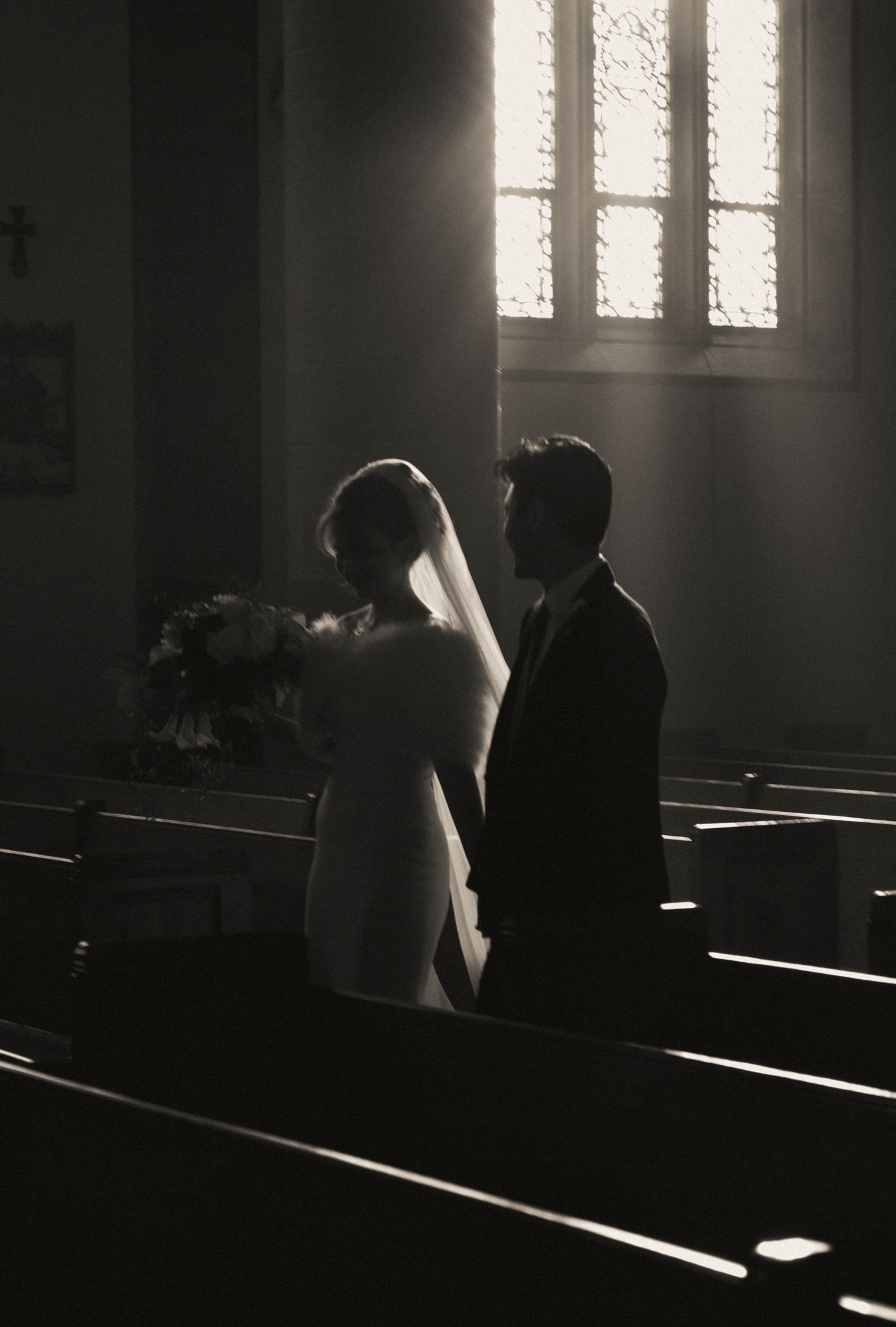 Silhouette of bride and groom standing in church pews with dramatic light streaming through window.