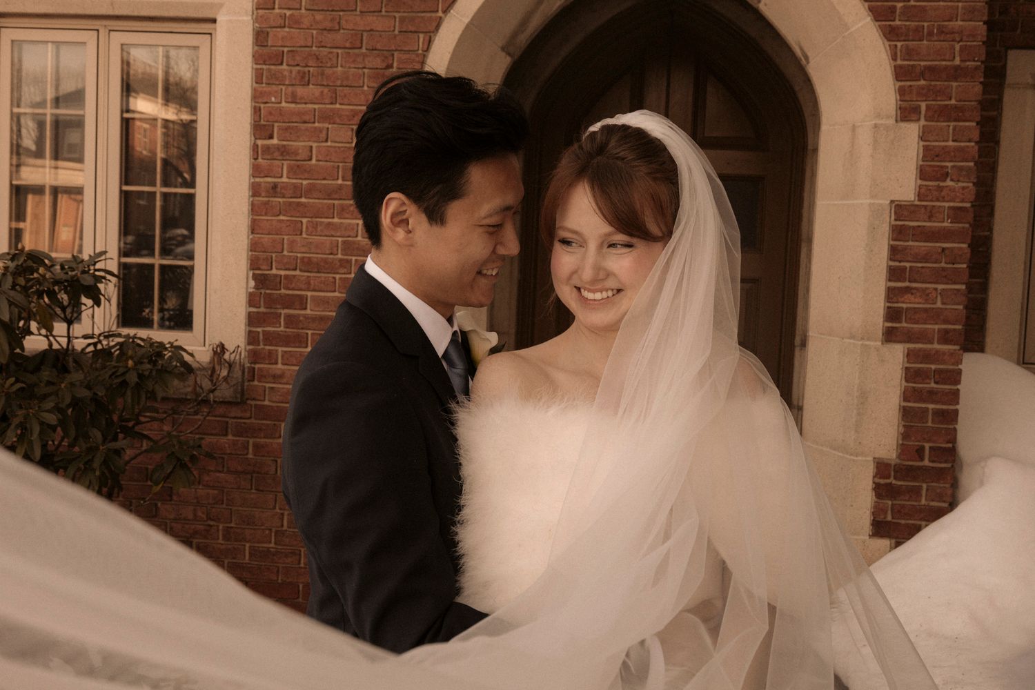 Newlywed couple sharing a tender moment, bride in white veil and gown with groom in black suit before brick building.
