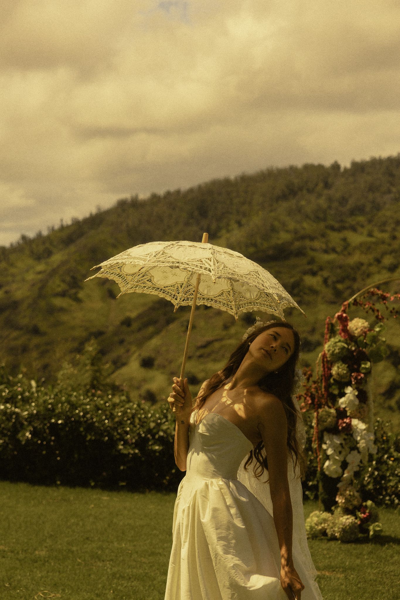 Woman in white dress holding lace parasol outdoors with green hills and cloudy sky backdrop.