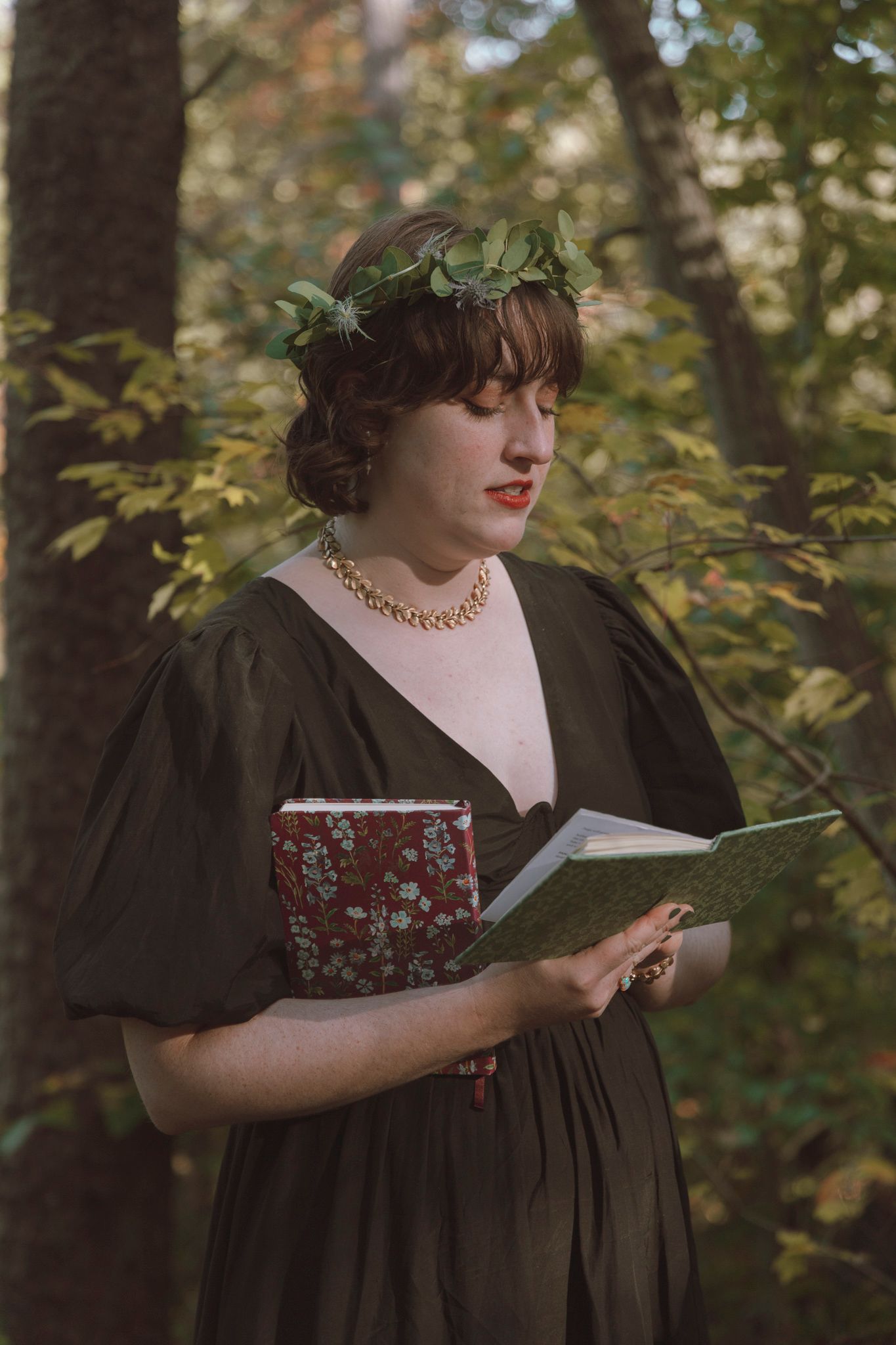 A person in a dark green dress and leaf crown stands in a forest setting while reading from a book.