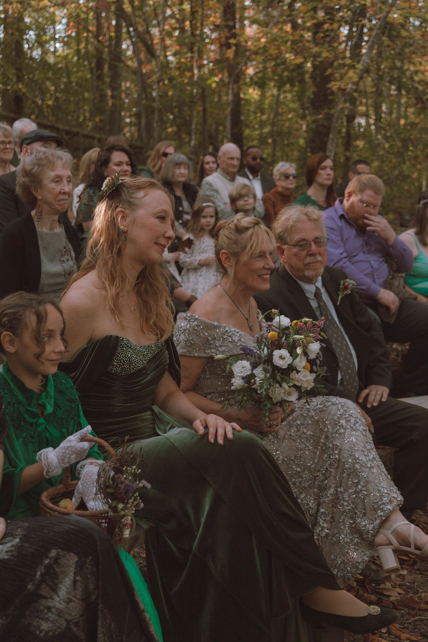 Wedding guests seated outdoors in autumn woods share joyful laughter during forest ceremony.