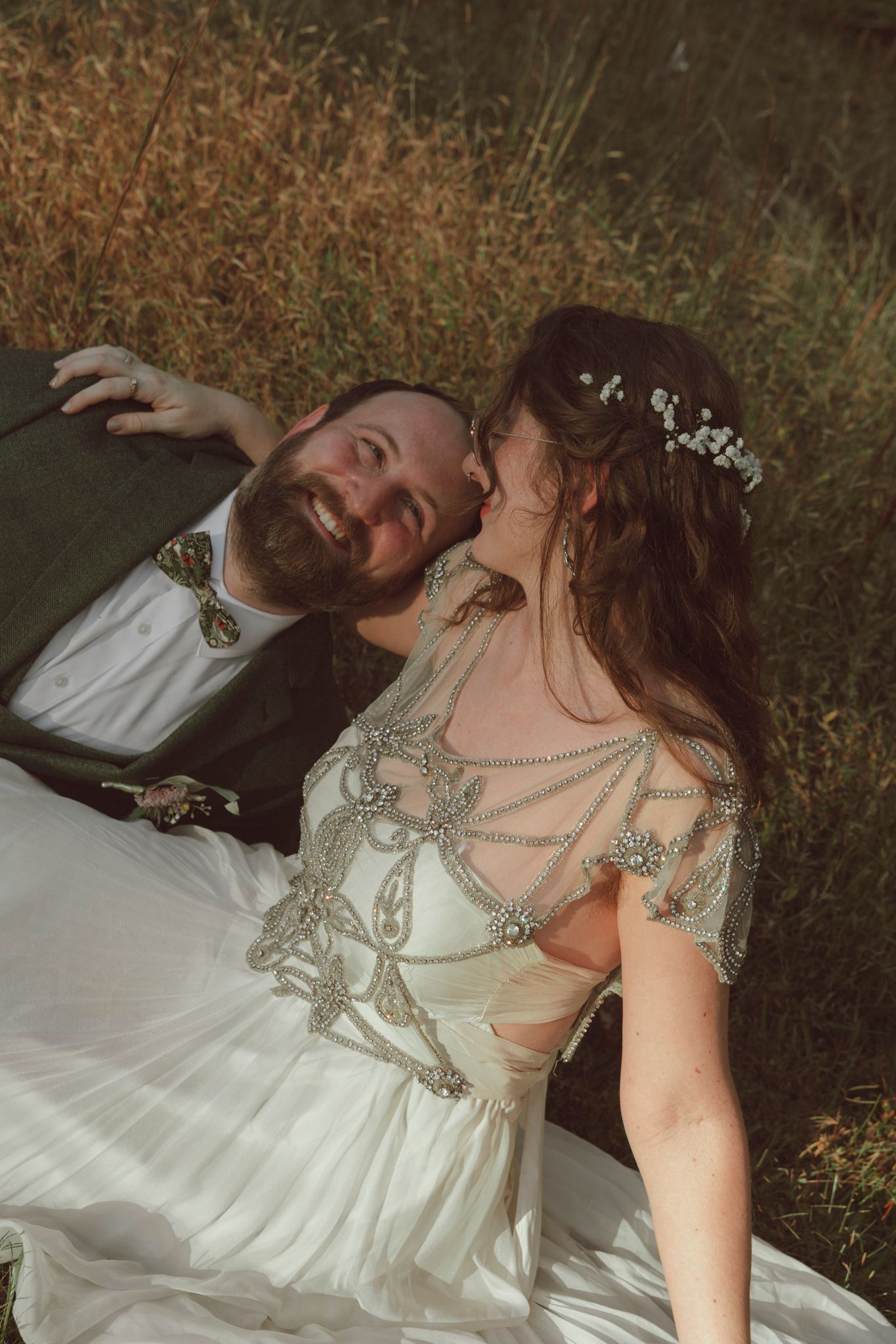 A romantic wedding photo shows a couple lying in autumn grass, with the bride wearing an ornate beaded dress and floral crown.