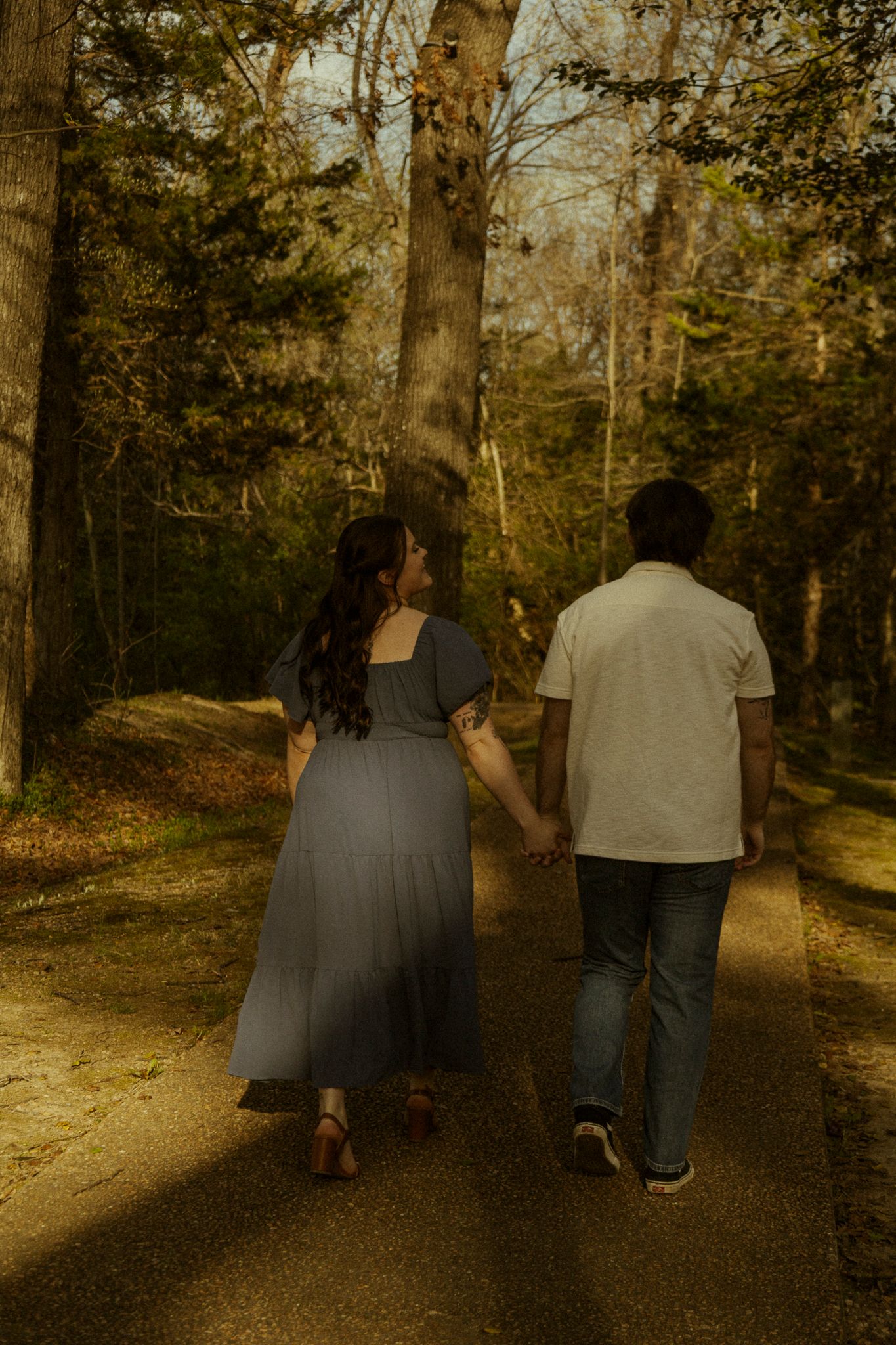 Couple holding hands walking down a sunlit forest path, woman in gray dress and man in white shirt and jeans.