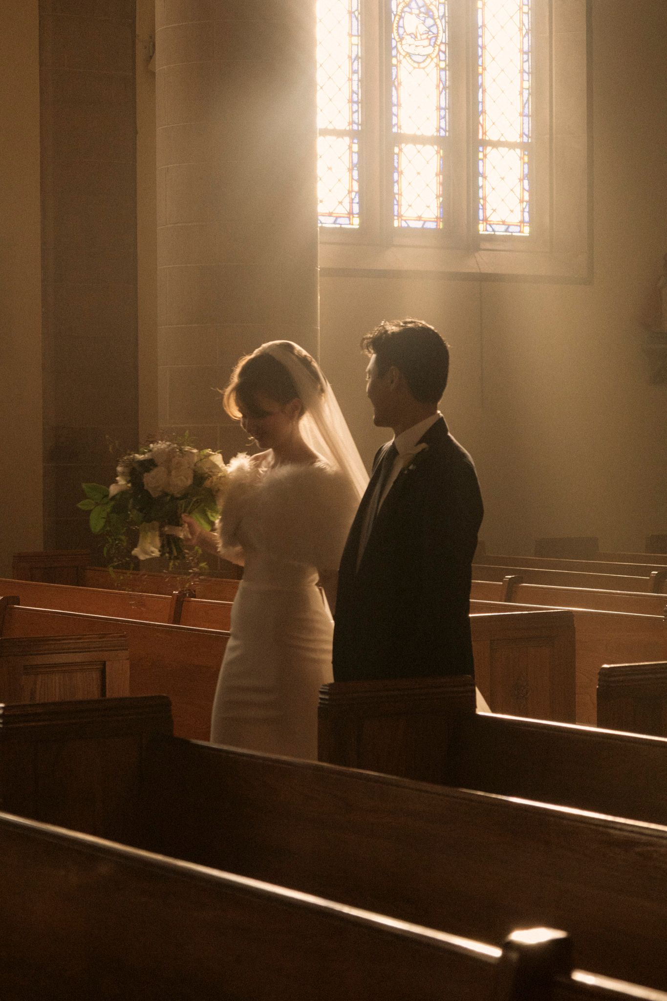 Bride and groom standing in sunlit church aisle with stained glass windows and wooden pews during wedding ceremony.