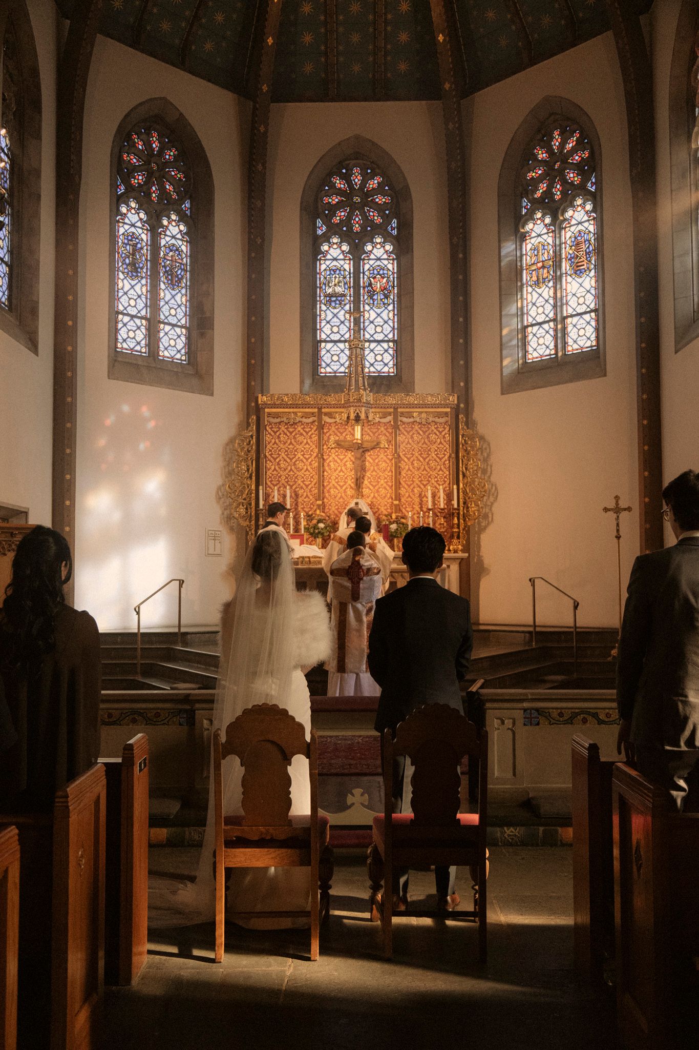 Bride in white veil and groom kneeling before ornate altar in church with beautiful stained glass windows.