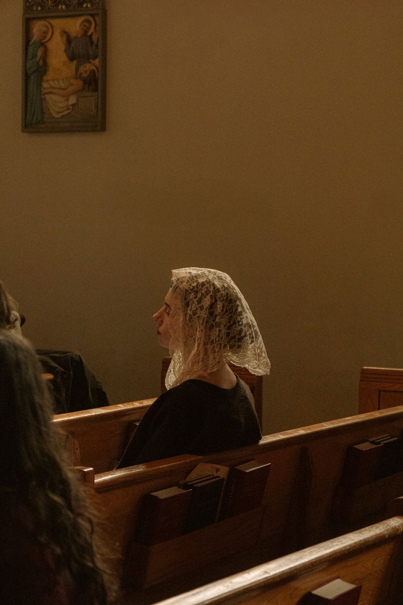 Elderly woman wearing white lace chapel veil sits in wooden church pew during wedding ceremony.