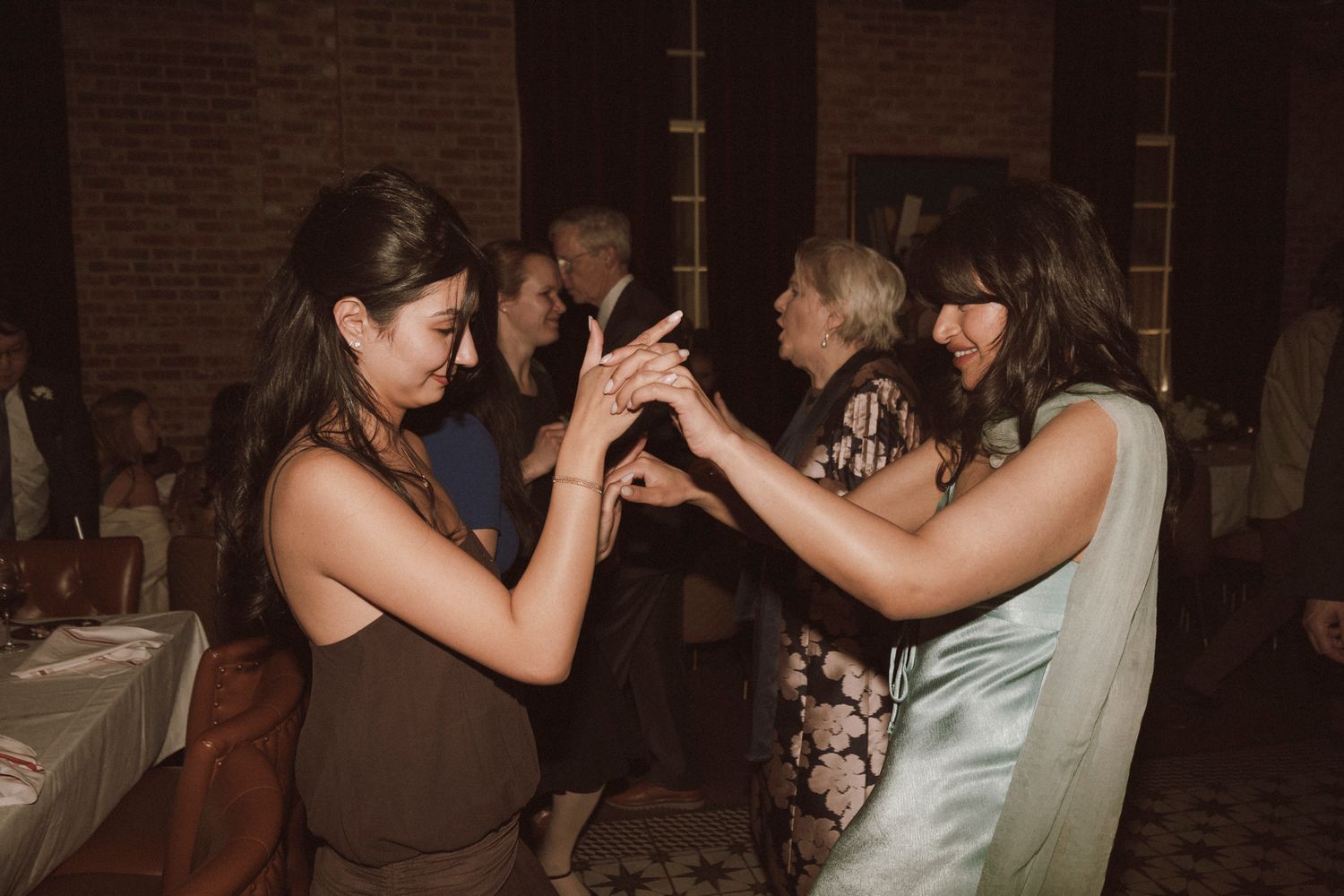 Women dancing together at a wedding reception in a dimly lit venue with brick walls.