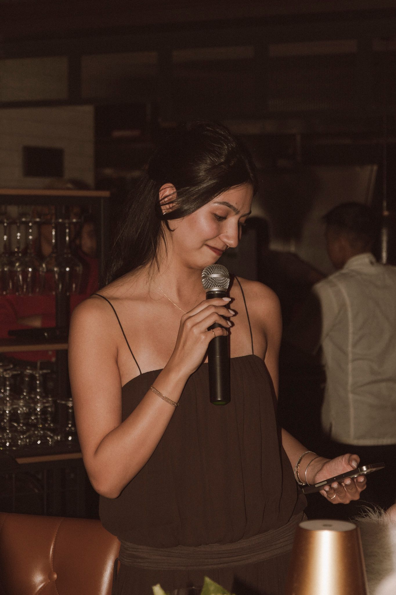 Young woman in brown dress speaking into microphone at an upscale restaurant event.