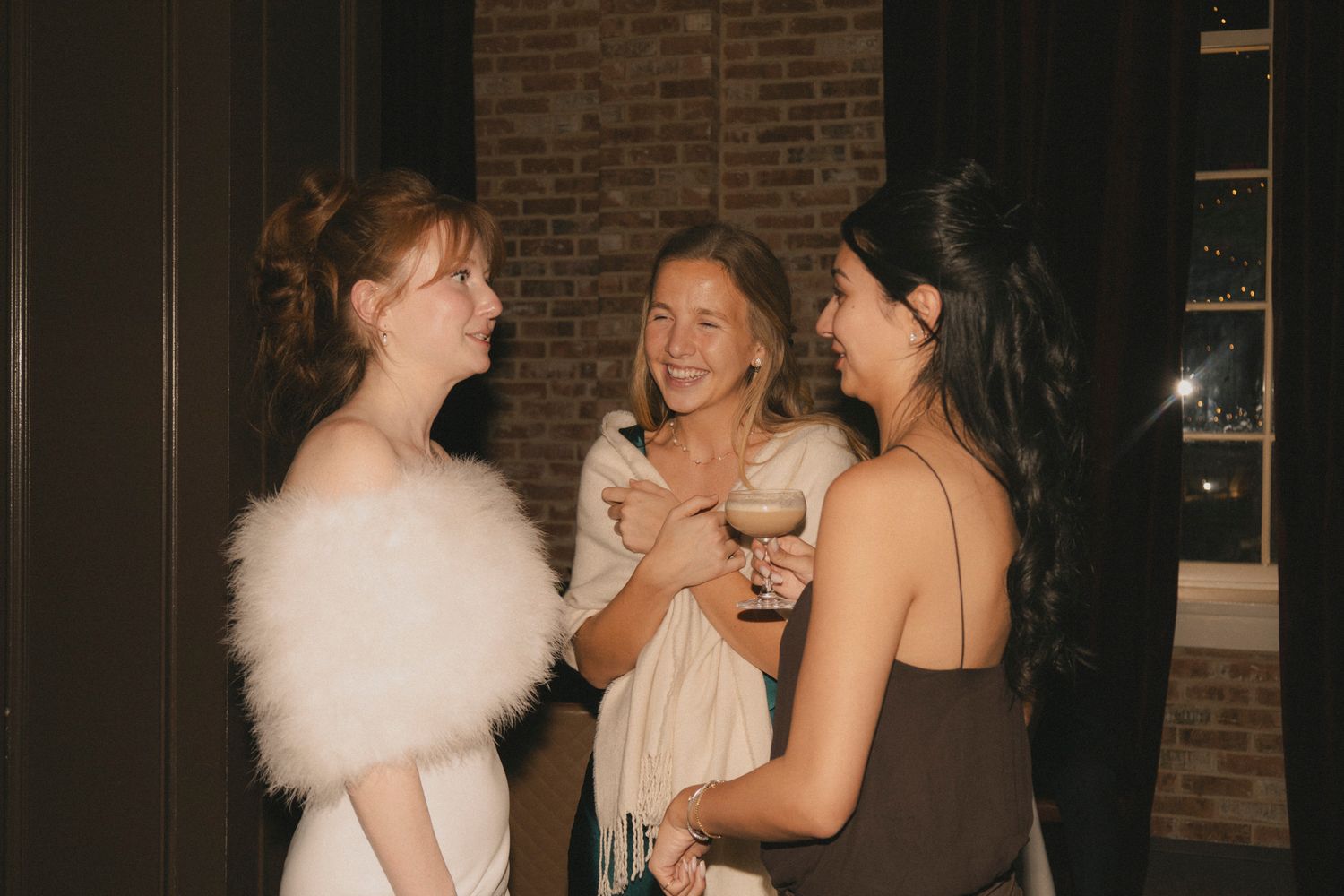 Bride in white fur stole laughs with two female wedding guests at an evening reception.