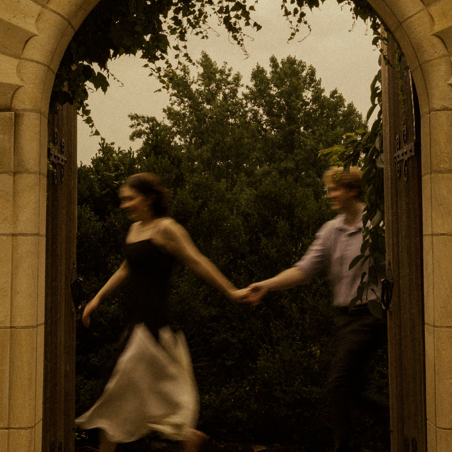 A blurred couple holds hands running through a stone archway surrounded by lush green trees in soft light.