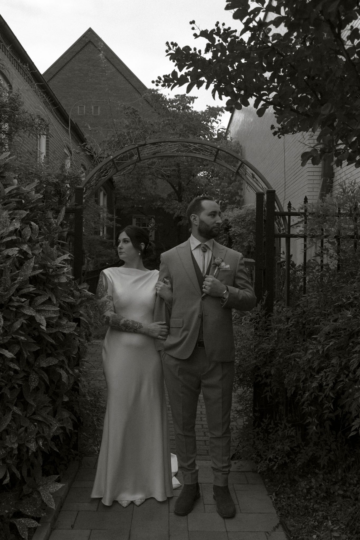 Artistic black and white wedding portrait of couple under garden archway surrounded by foliage.