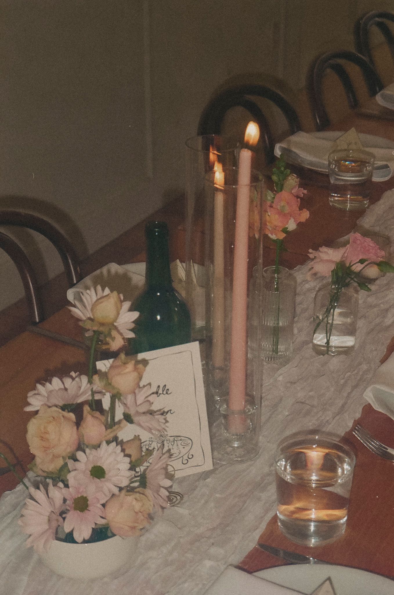 A romantic table setting with pink flowers, lit candles, and wine bottles creates an intimate dining atmosphere.