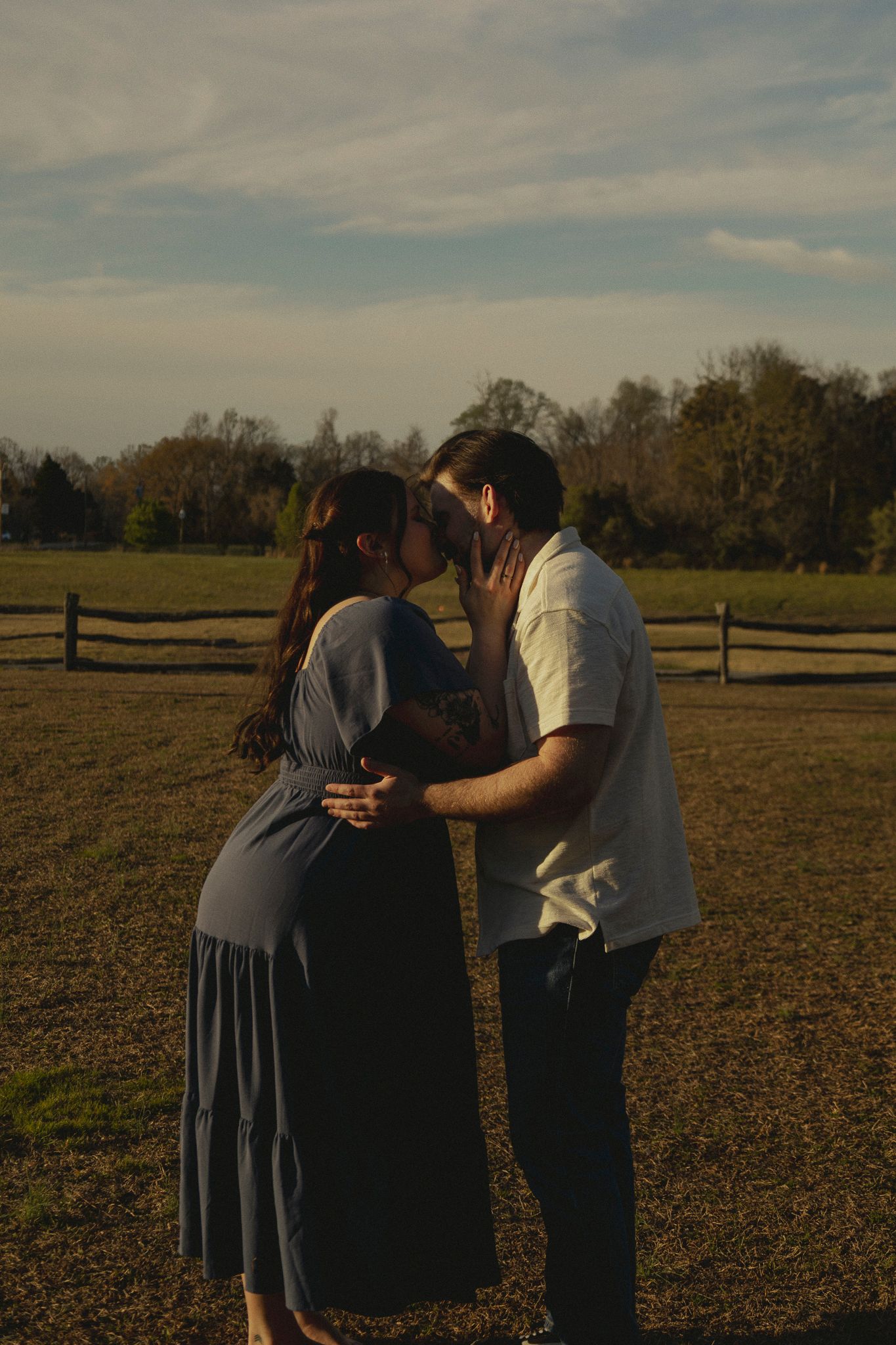 A couple shares an intimate moment touching foreheads in a golden field at sunset, the woman in a blue dress embraced tenderly.