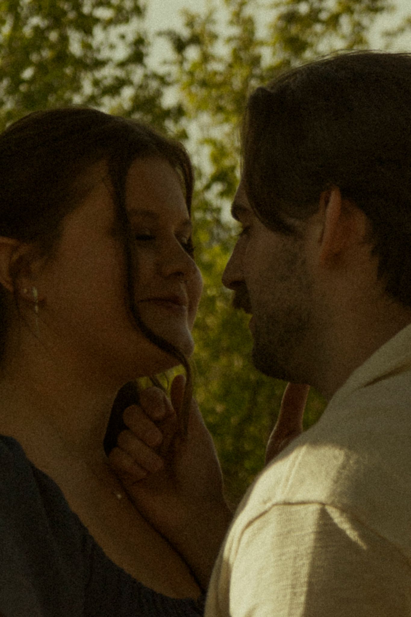 A couple shares an intimate moment outdoors, faces close together, surrounded by lush green trees in warm golden light.