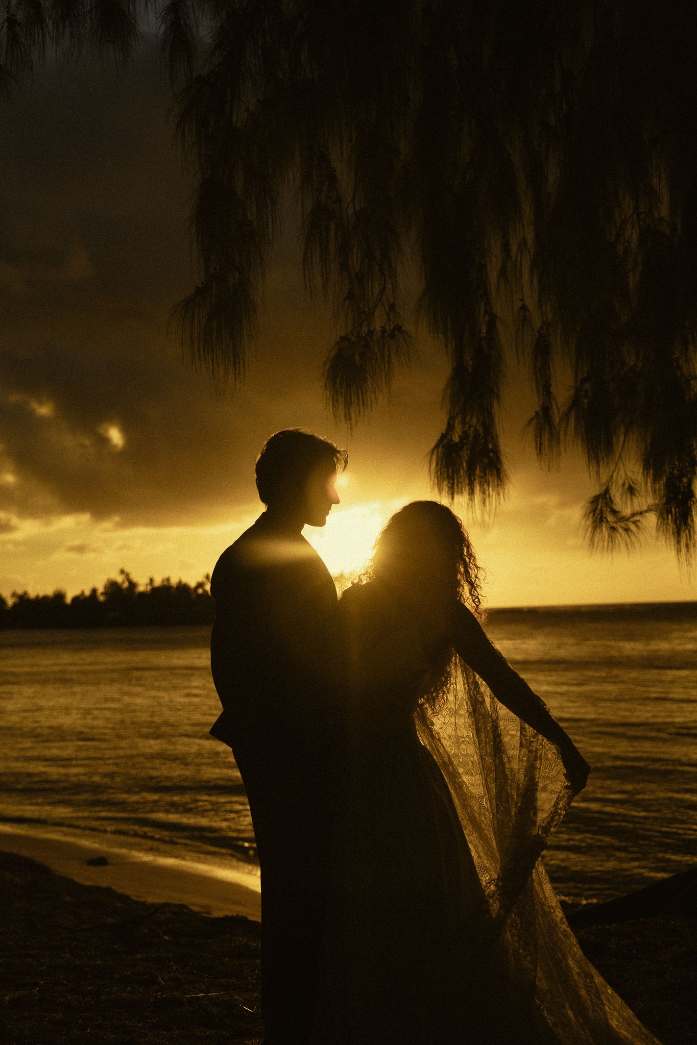 Silhouette of a bride and groom embracing at sunset on a tropical beach, with golden sky and ocean backdrop.