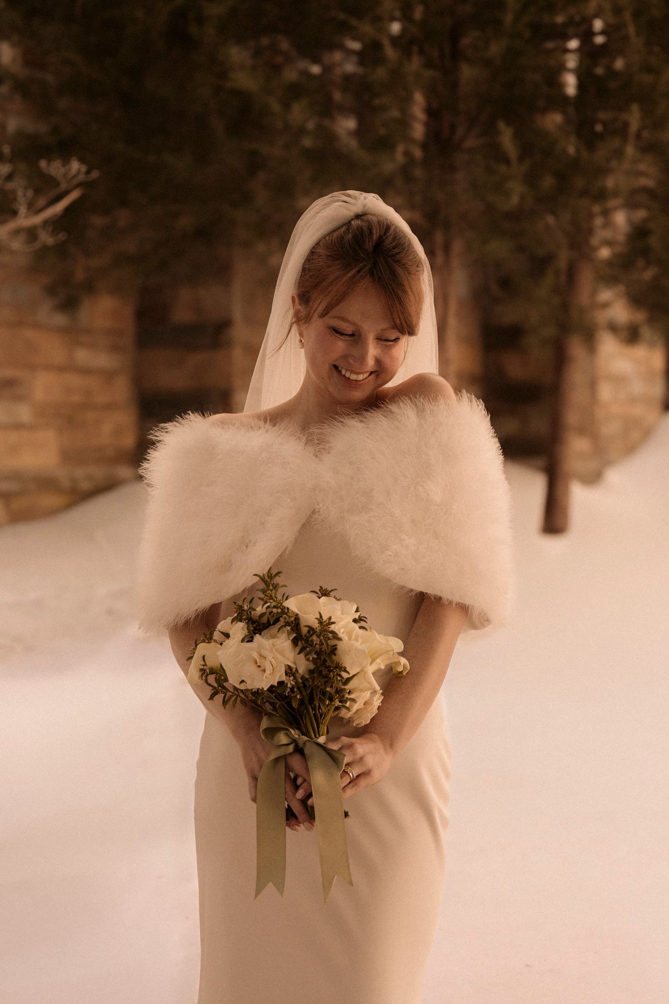 Smiling bride in white gown, fur shawl, and veil holds bouquet in snowy winter forest setting.