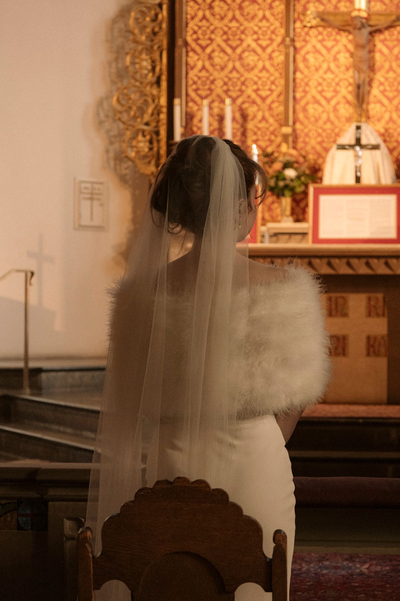 Bride in white gown and veil kneeling in prayer before ornate church altar.