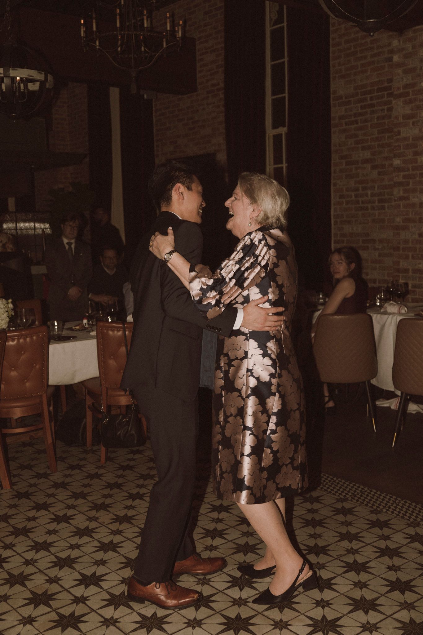 Groom dancing with his mother in a floral dress on the patterned tile floor at wedding reception.