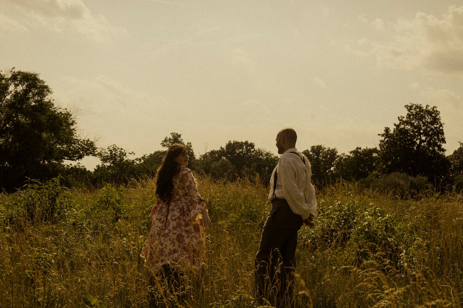 A couple stands in a lush green meadow at golden hour, surrounded by tall grass and trees in the background.