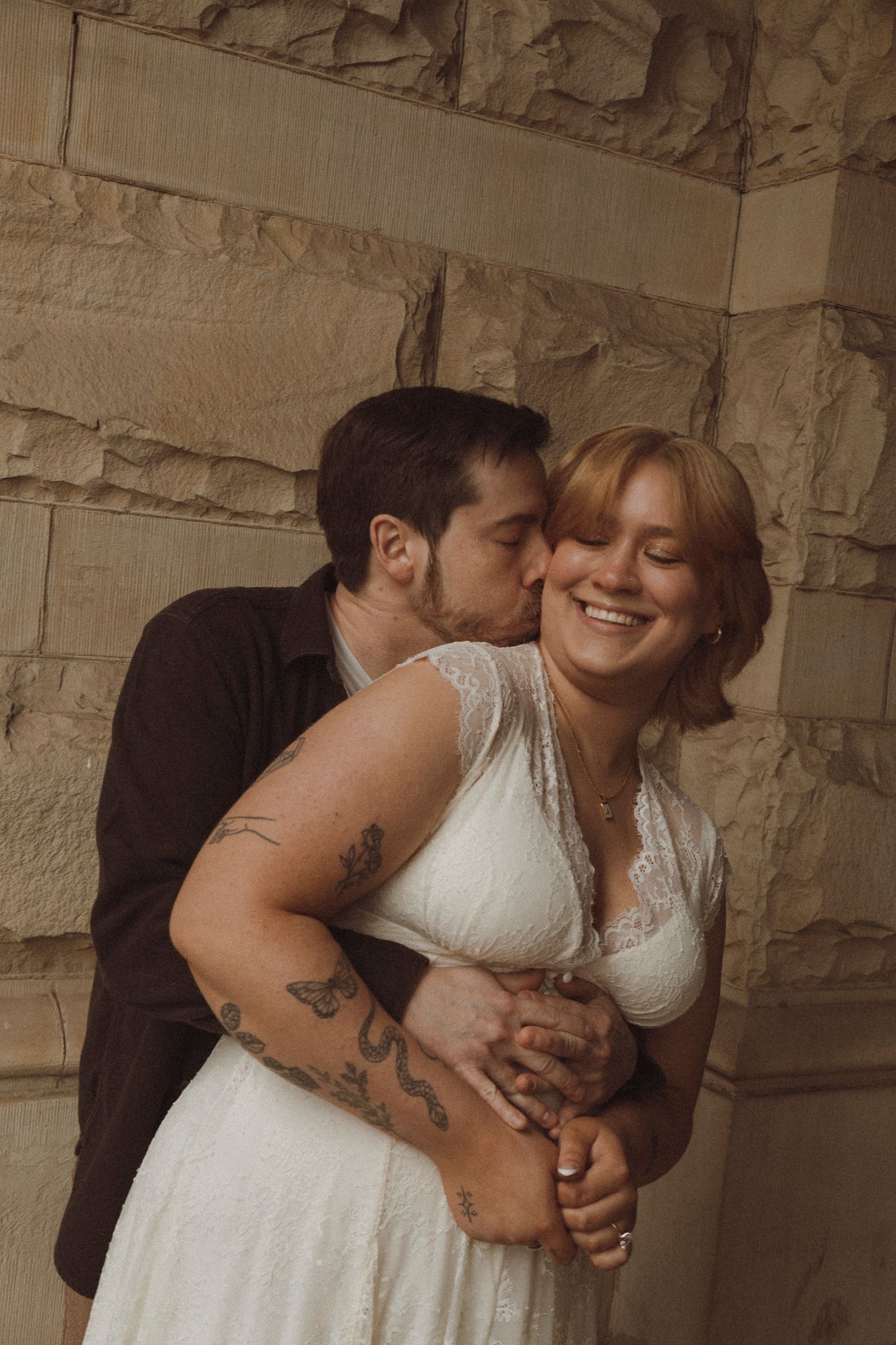 A romantic couple embracing in casual attire against a stone wall background.
