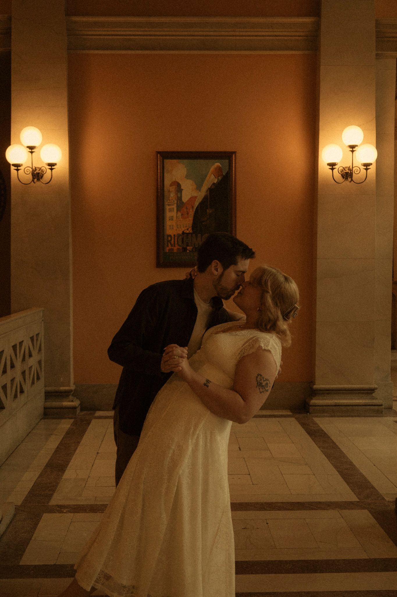 A couple in formal wedding attire share a romantic dance in an elegant hallway with wall sconces and marble floors.