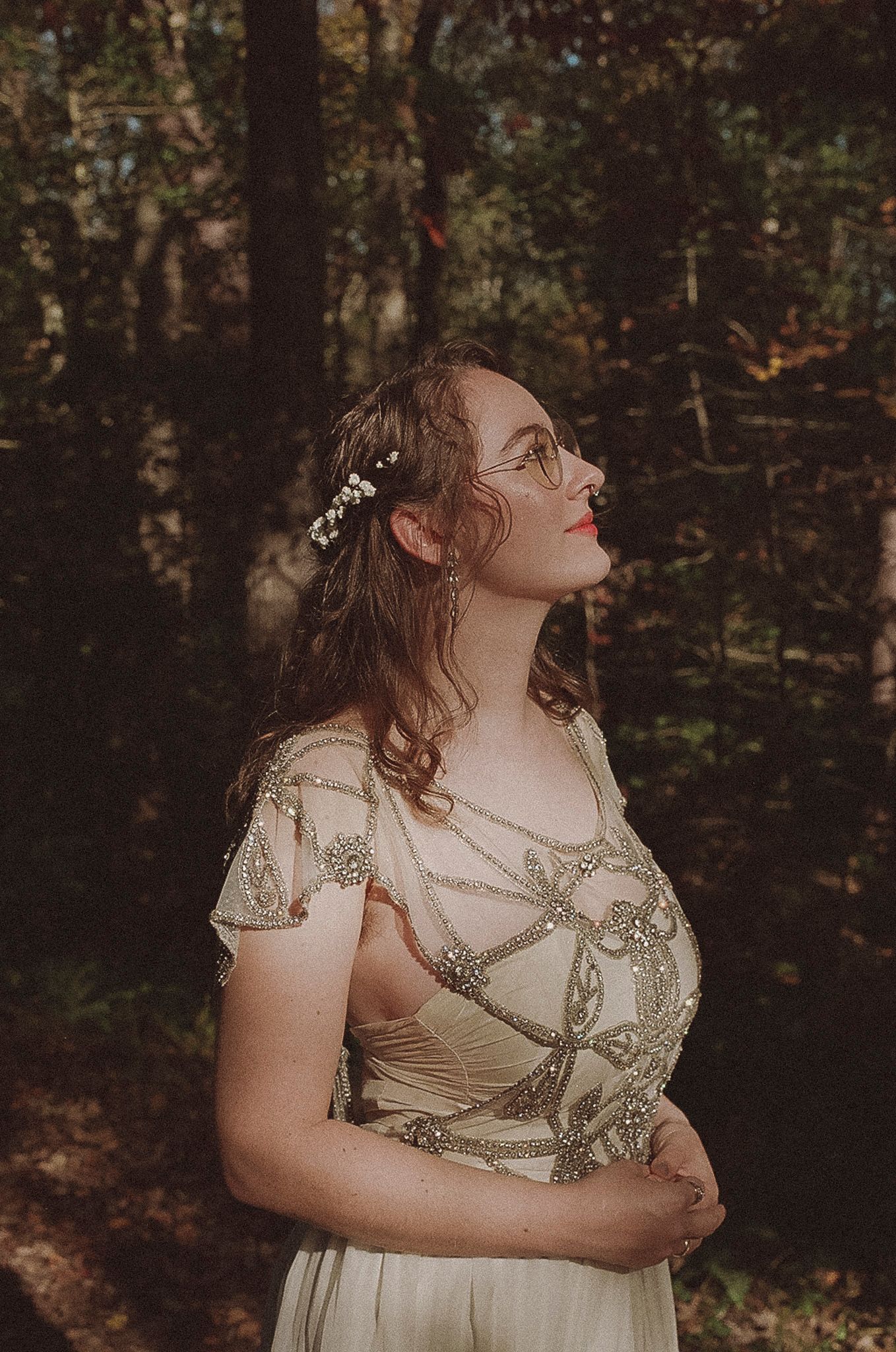 A dreamy woodland portrait shows a woman in a vintage-style beaded dress with flowers in her hair looking upward.
