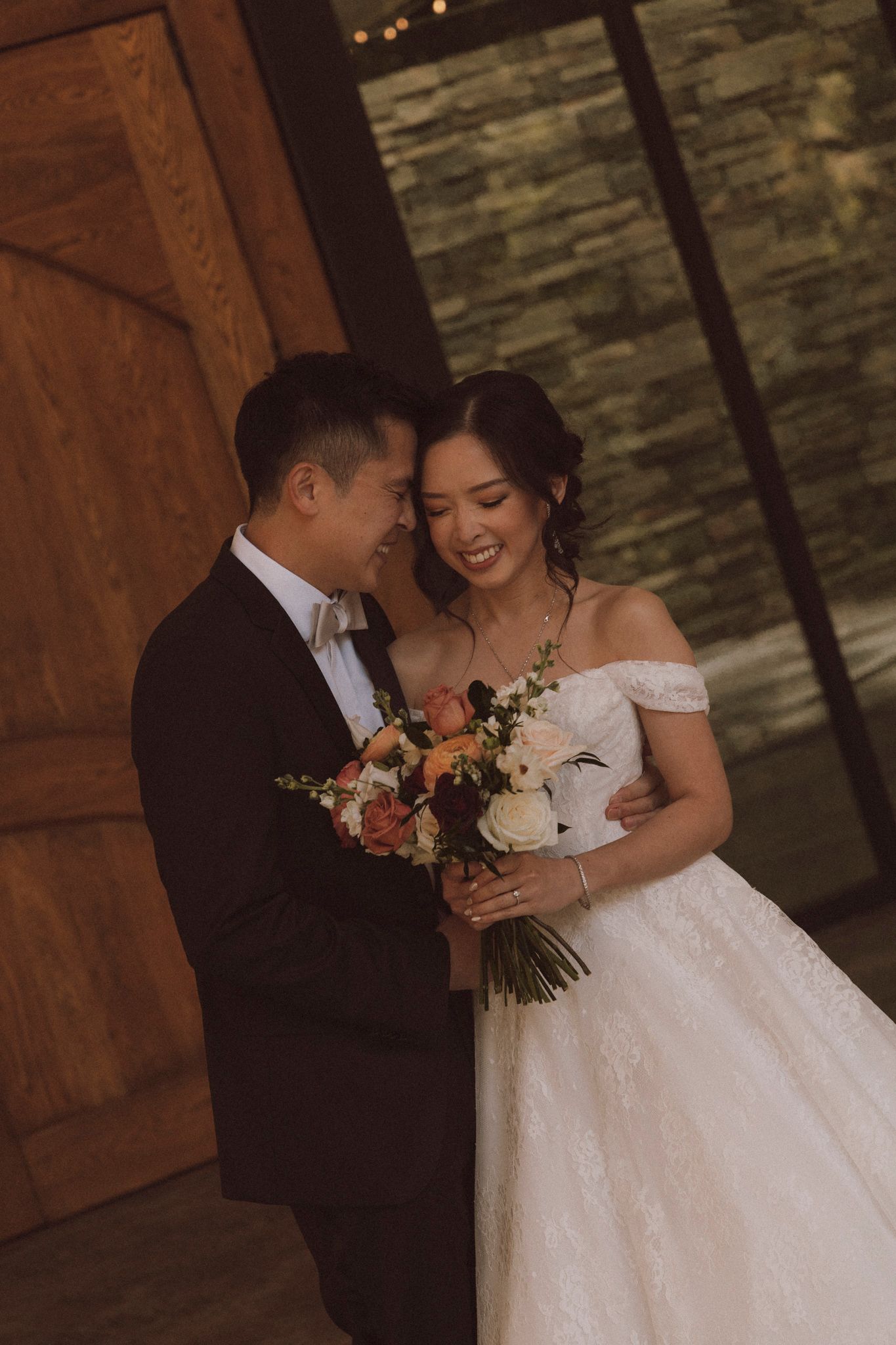A romantic wedding moment with a couple sharing an intimate pose against a stone wall backdrop, holding a fall-toned bouquet.