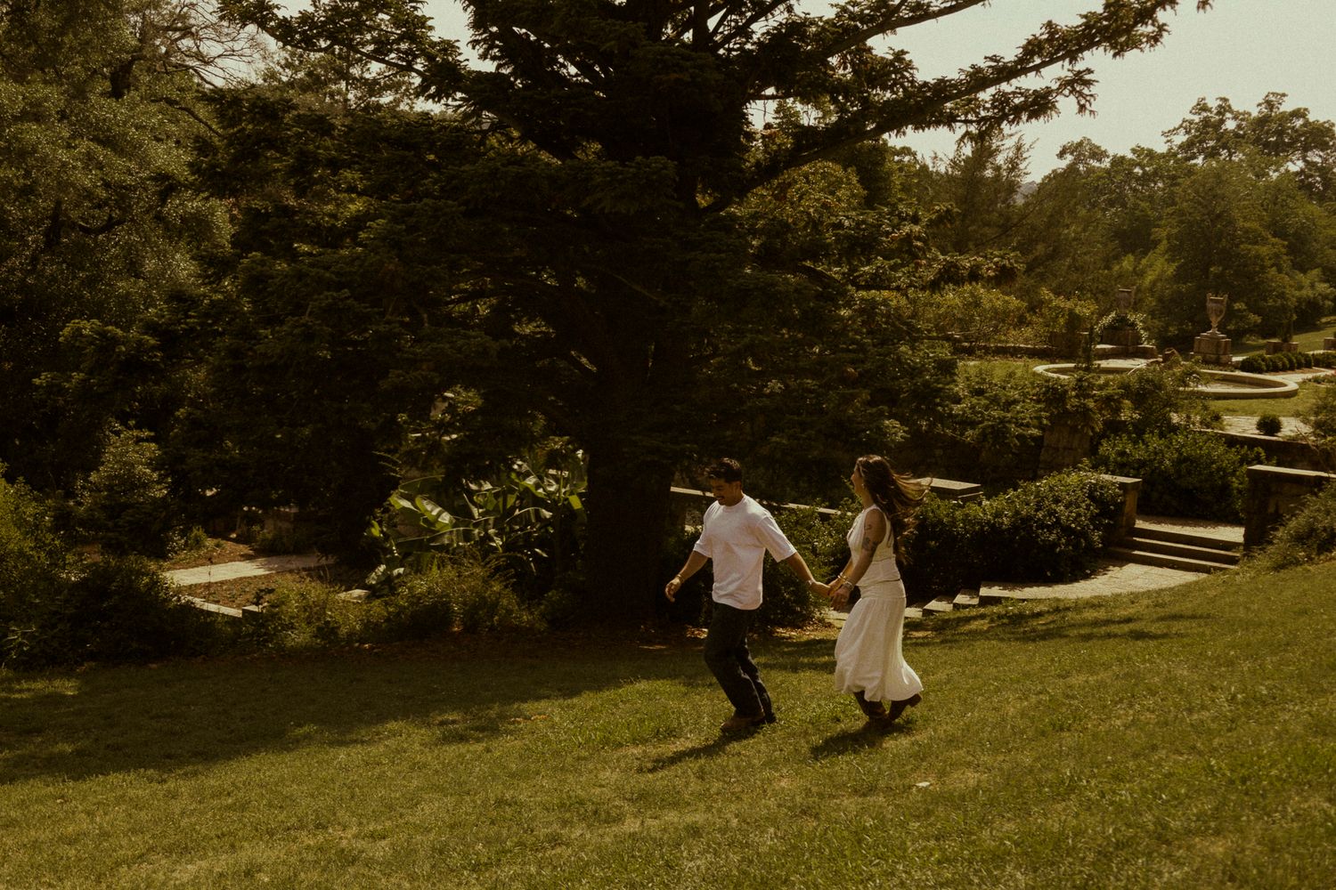 Couple holding hands running through lush green garden grounds on a sunny day.