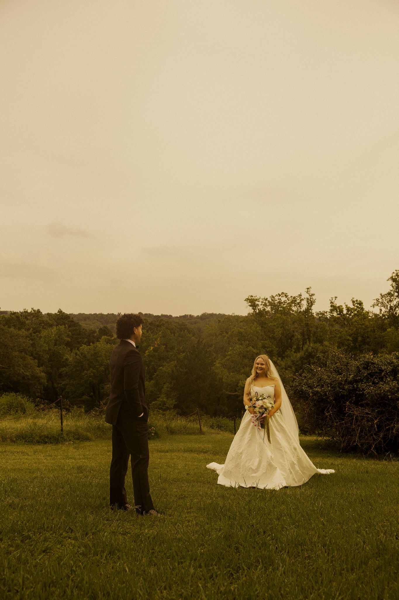 Bride and groom share a romantic first look on a lush green lawn at dusk, with rolling wooded hills behind them.