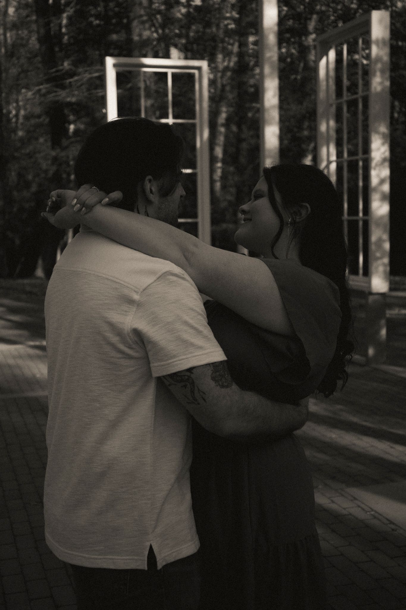 Black and white photo of couple embracing tenderly outdoors near decorative window frames.