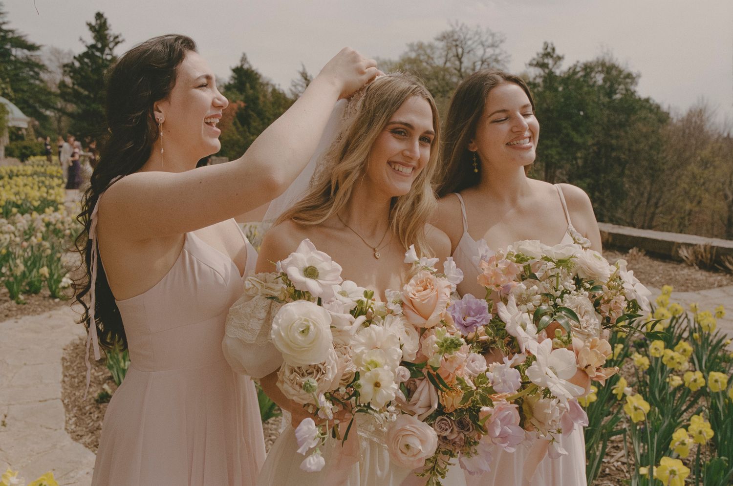 Wedding party wearing blush pink dresses holds bouquets while posing together outdoors in natural sunlight.