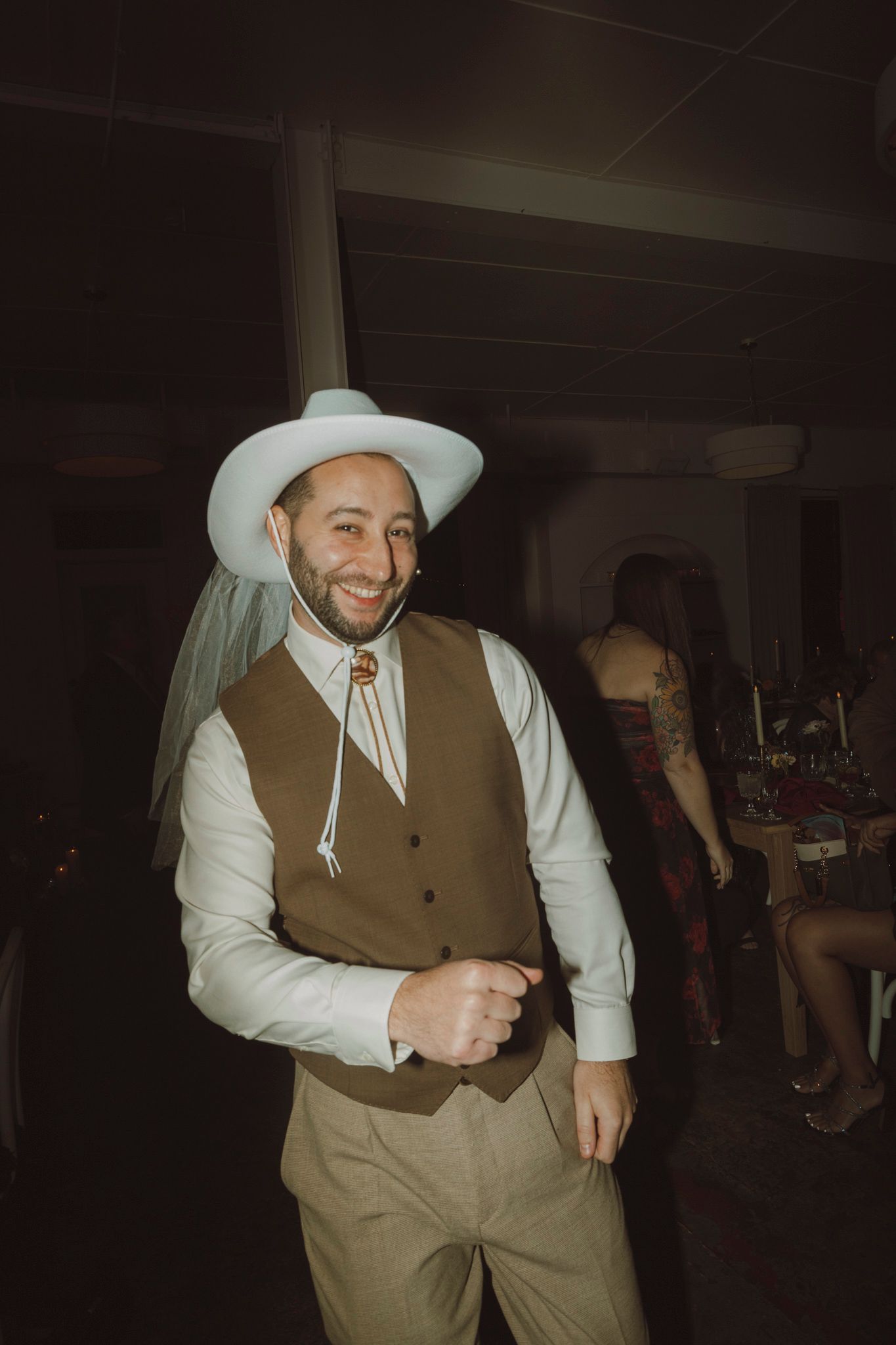 A person in western attire with a tan vest and white cowboy hat dances and moves across a dark room.