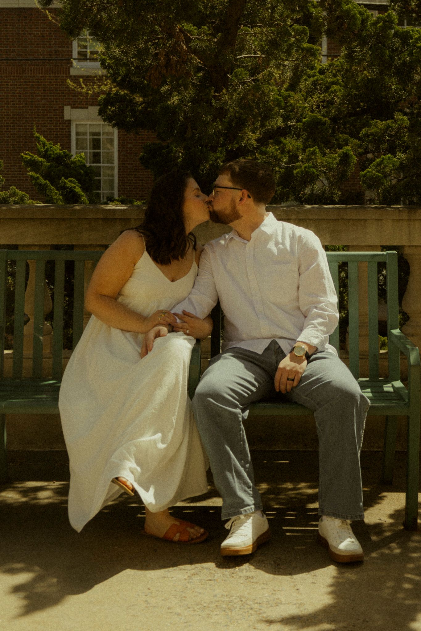 A couple shares a romantic kiss while sitting on a green bench outdoors, wearing wedding attire.
