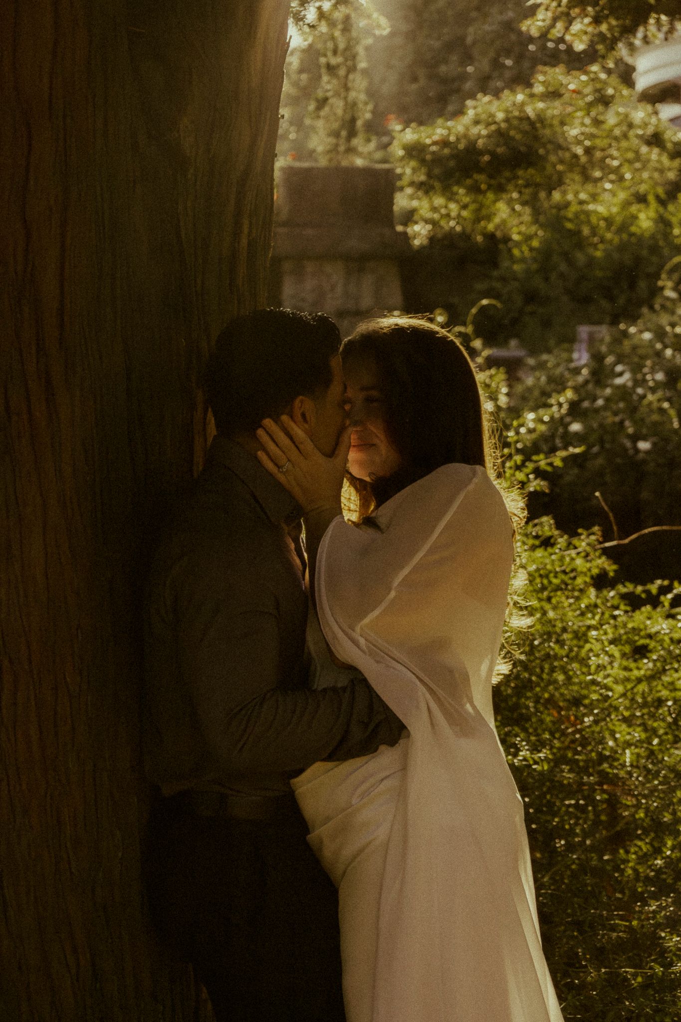 Couple sharing a romantic moment leaning against a large tree in a sunlit garden, bride in white dress.
