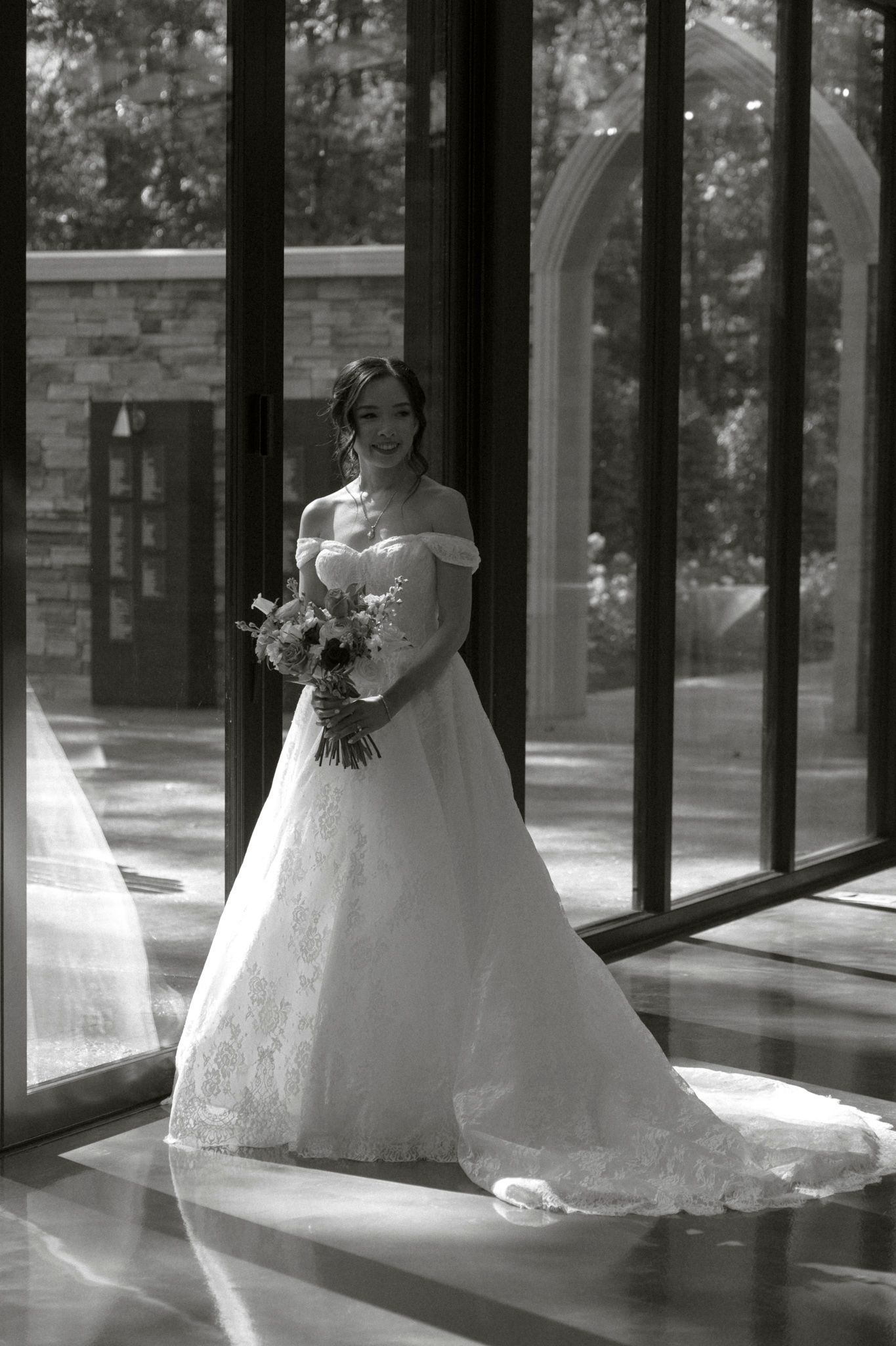 A black and white panoramic photo shows a bride in a flowing white gown standing in a sunlit corridor with glass windows.