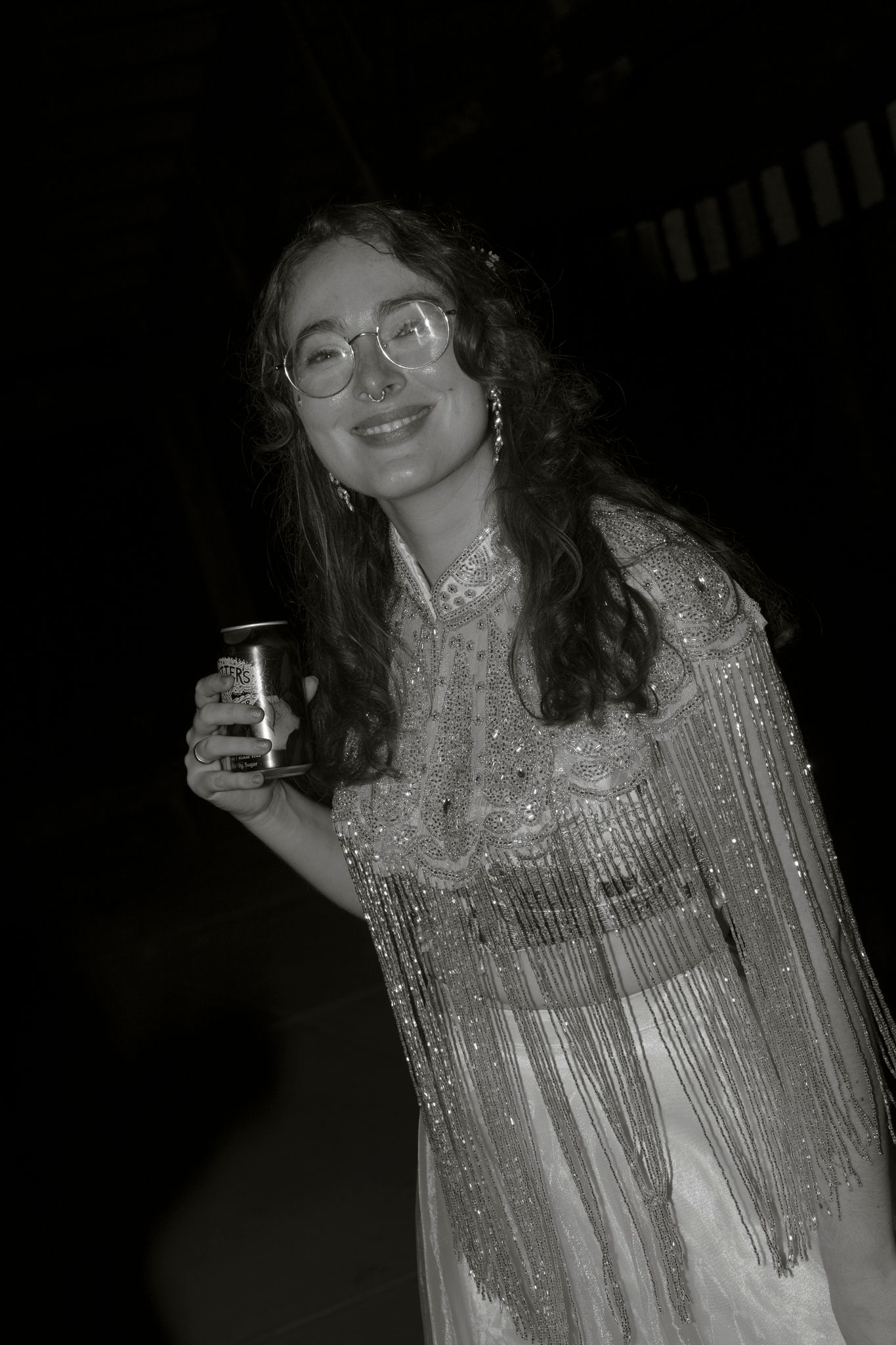 A person wearing a sparkly silver fringe dress poses with a drink in a dark setting.