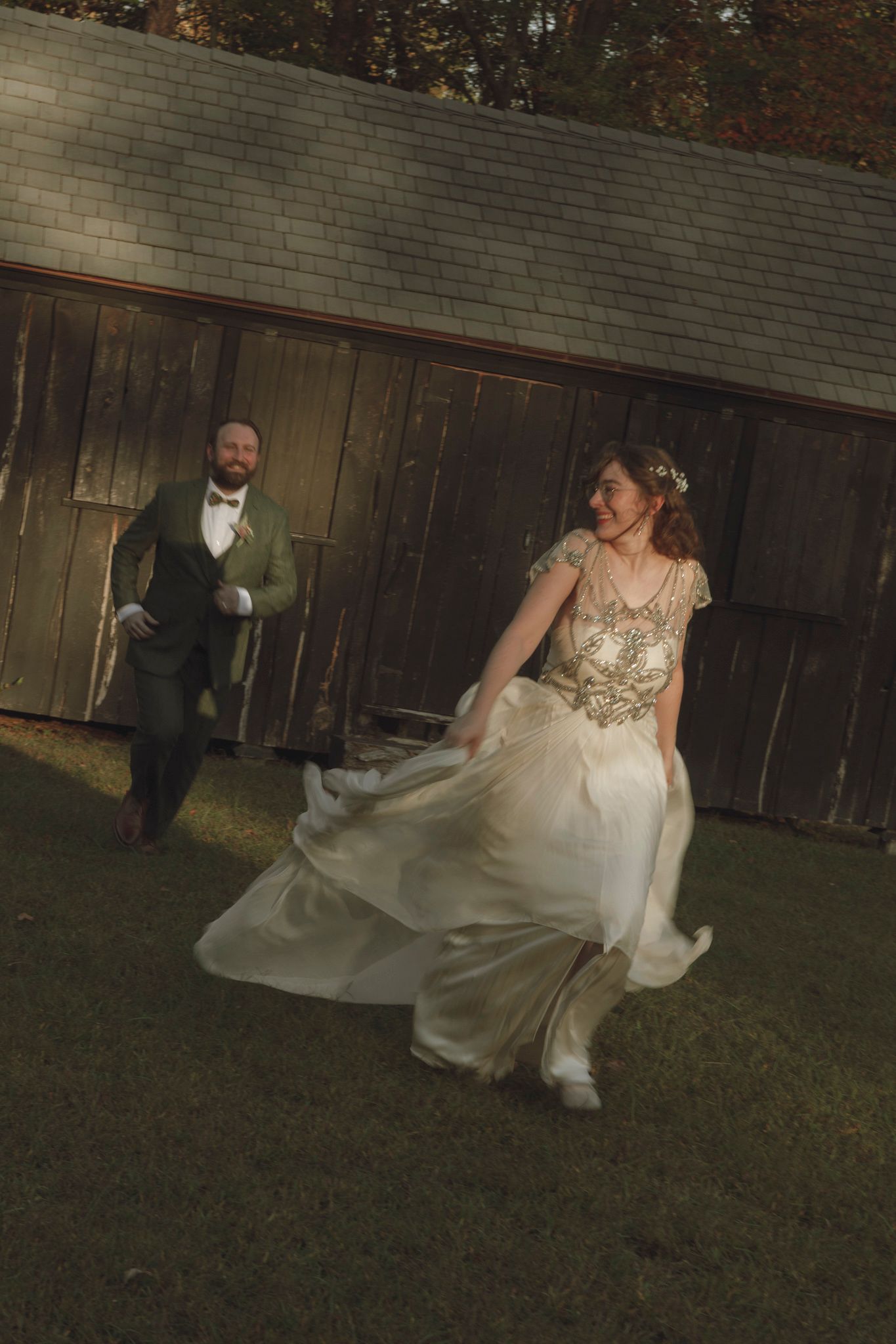 A couple in formal wedding attire dances and twirls together on a lawn at dusk with a wooden fence in the background.