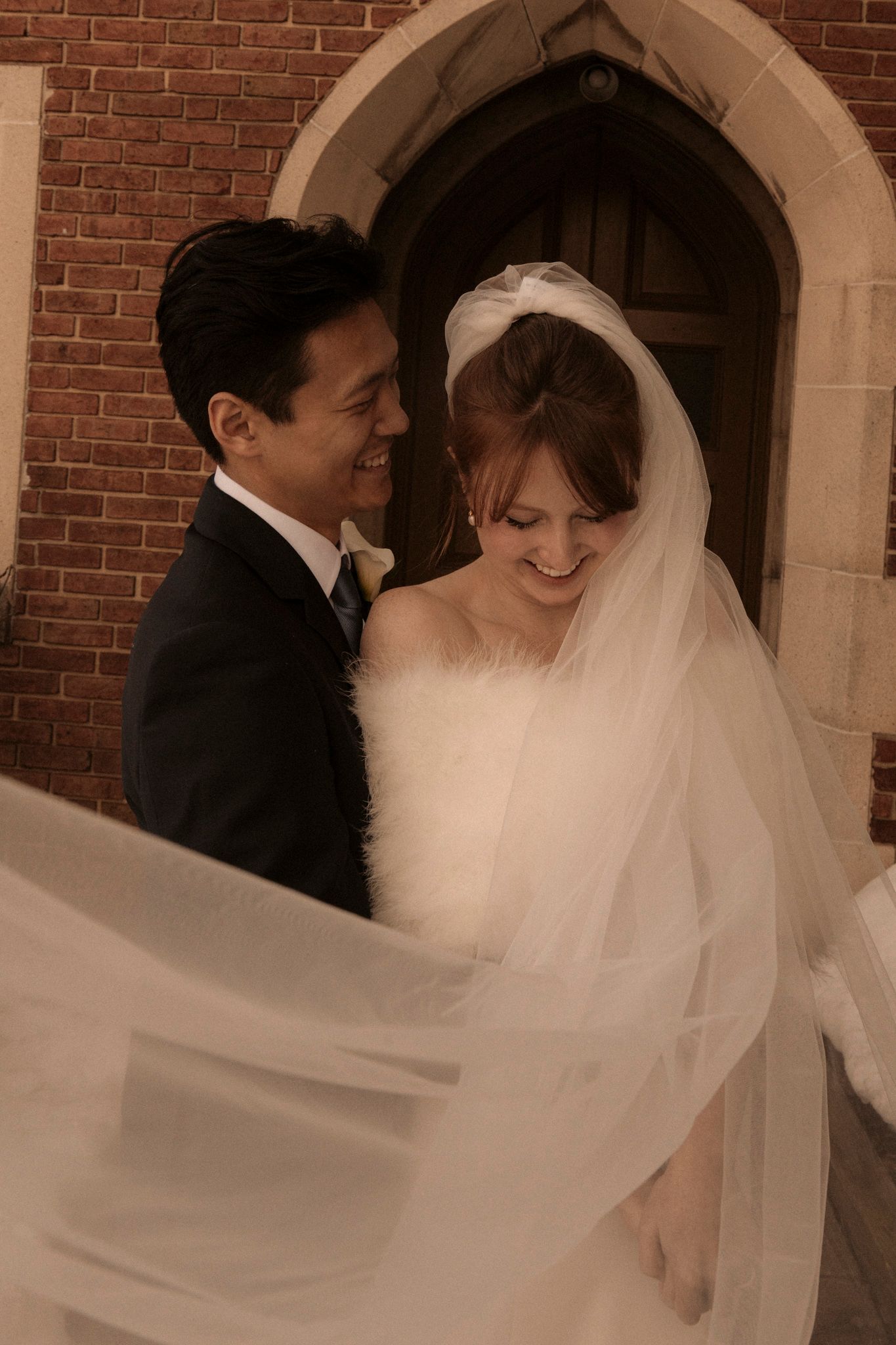 Smiling newlyweds pose outside a brick church doorway as the bride's veil flows in the wind.
