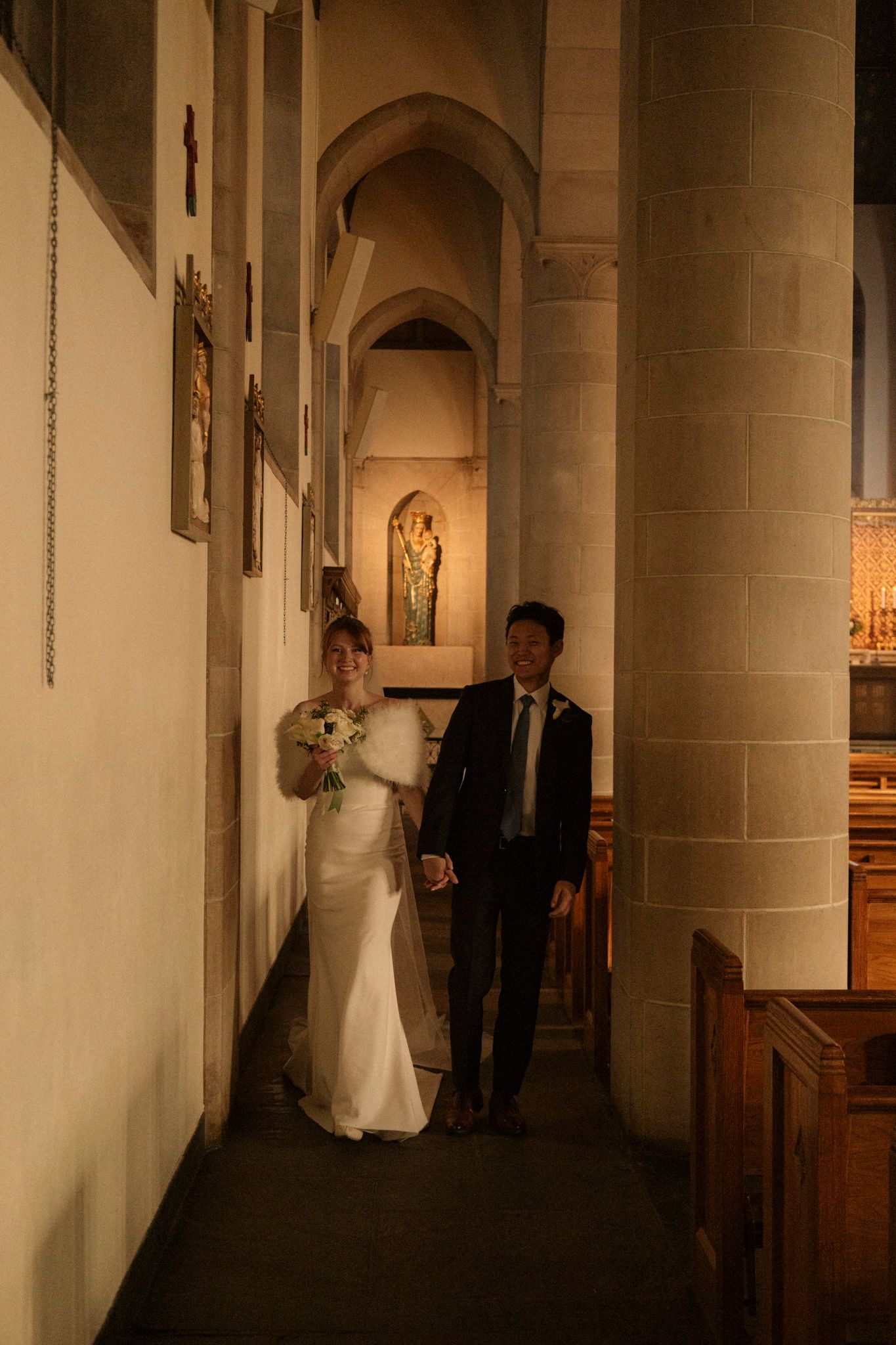 Bride and groom walk hand in hand through a dimly lit stone church corridor during their wedding.