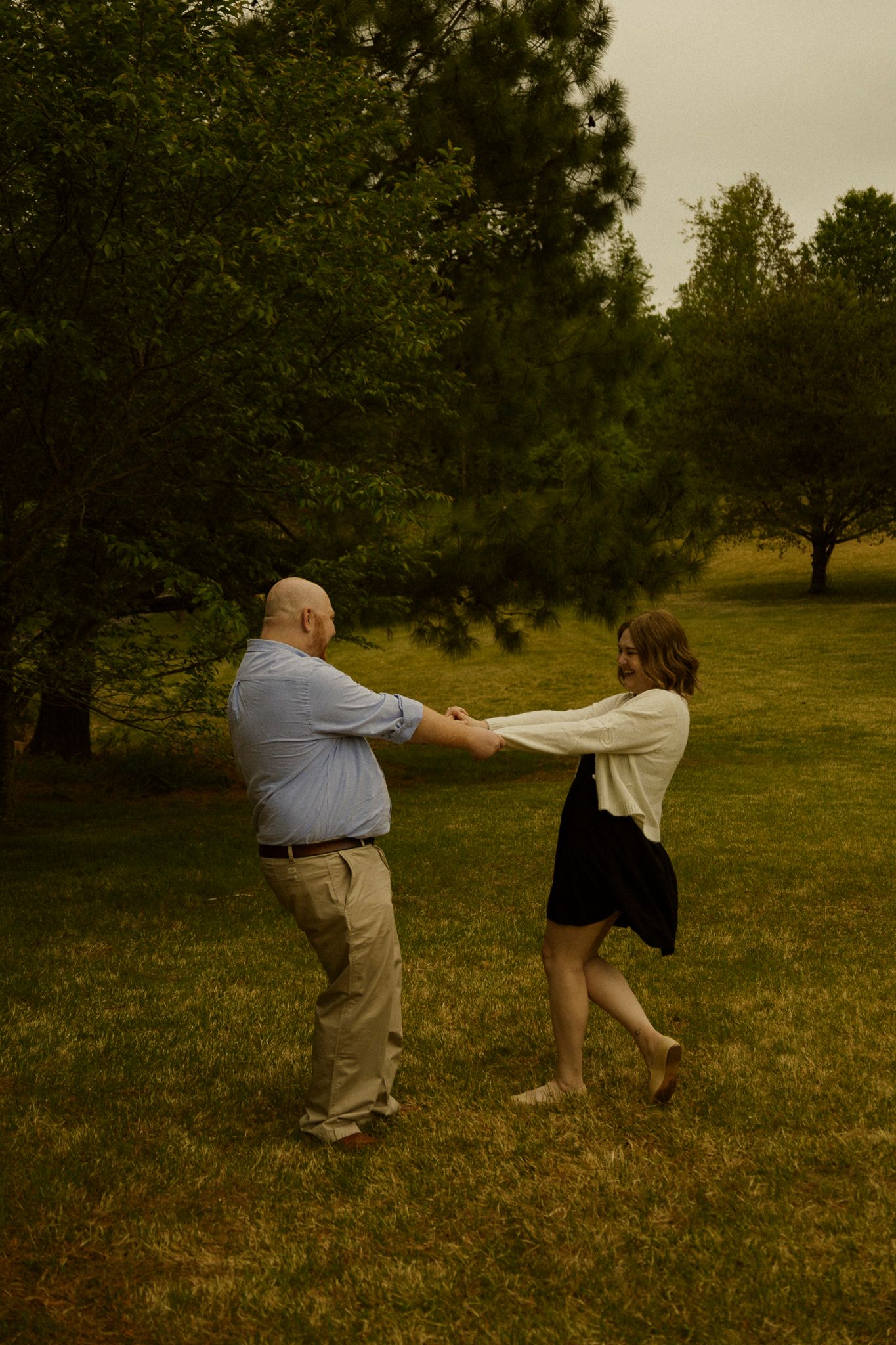 A couple dances and spins together on a green lawn surrounded by lush trees in a park setting.