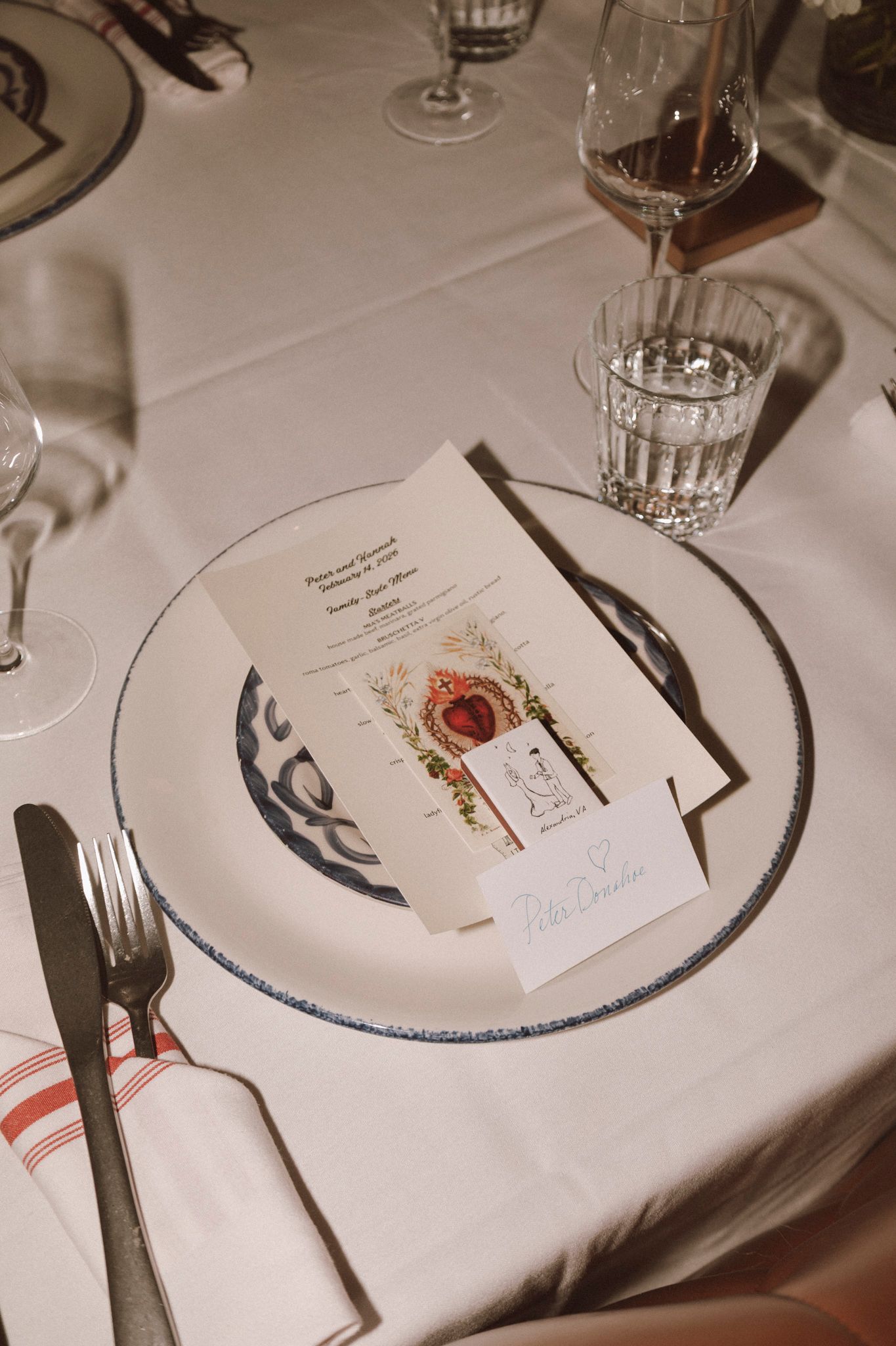 Elegant wedding table setting with blue-rimmed charger plate, menu card, place card, crystal glasses, and silverware on white linen.