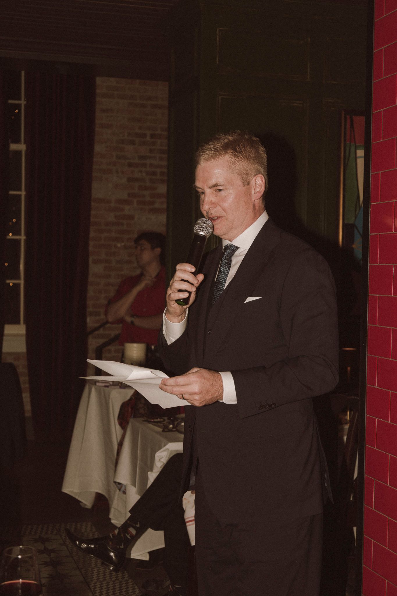 Elegant man in black suit speaks into microphone while reading from papers at a formal event.