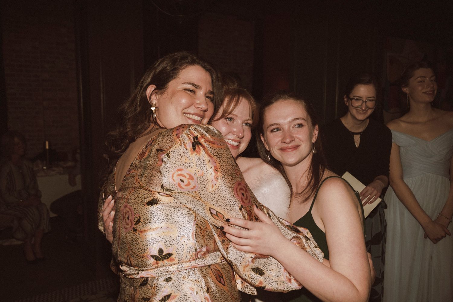 Three women in formal attire sharing a warm group hug at a wedding reception celebration.