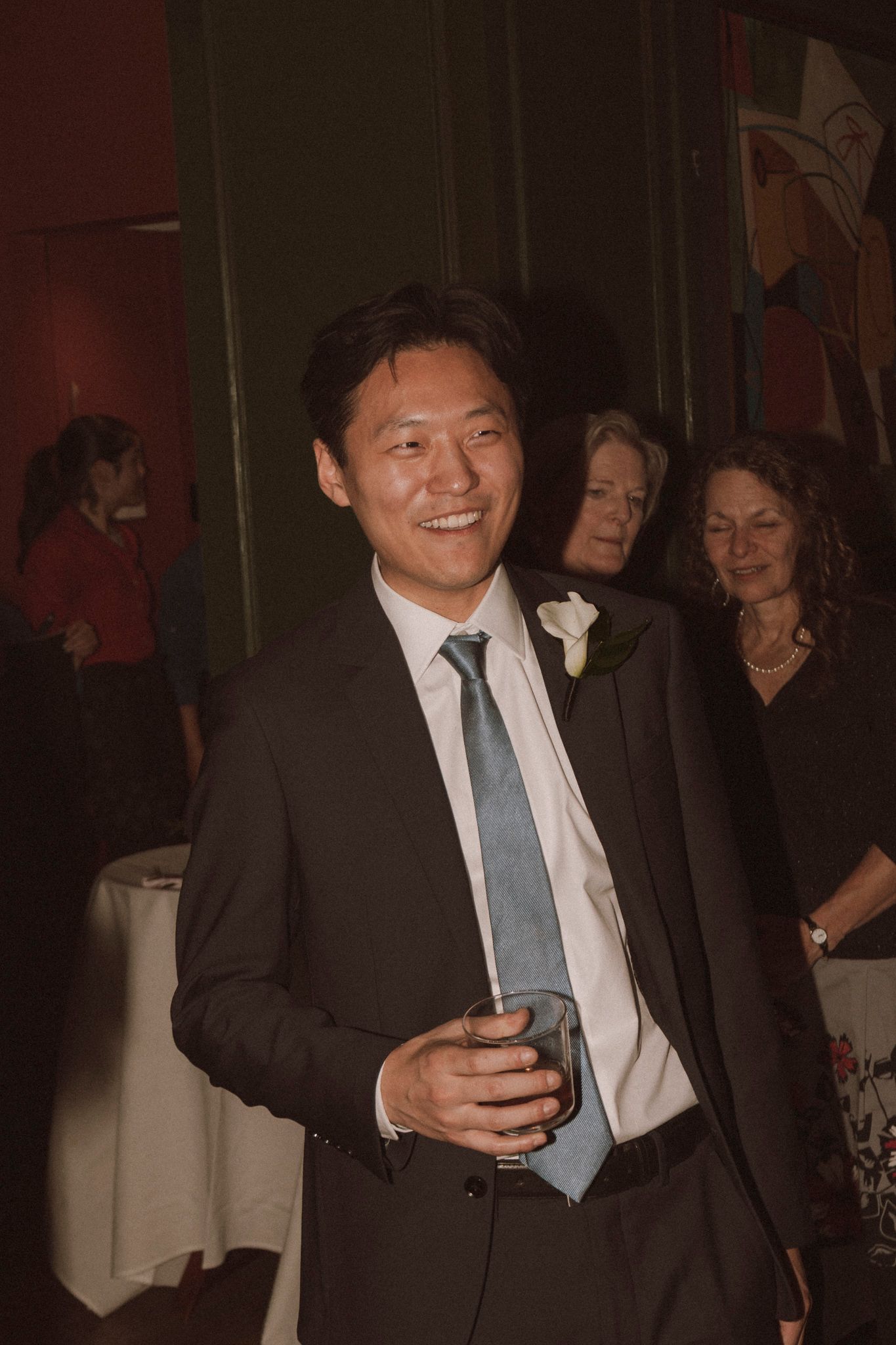 A smiling man in a dark suit and blue tie holds a drink at a formal wedding reception.
