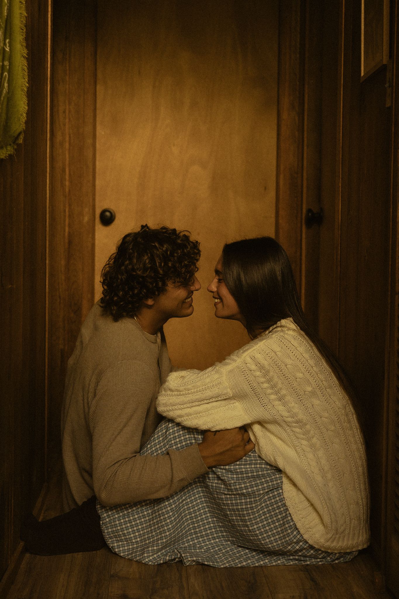 A couple sits intimately on the floor against a wooden door, gazing at each other in warm, dim lighting.