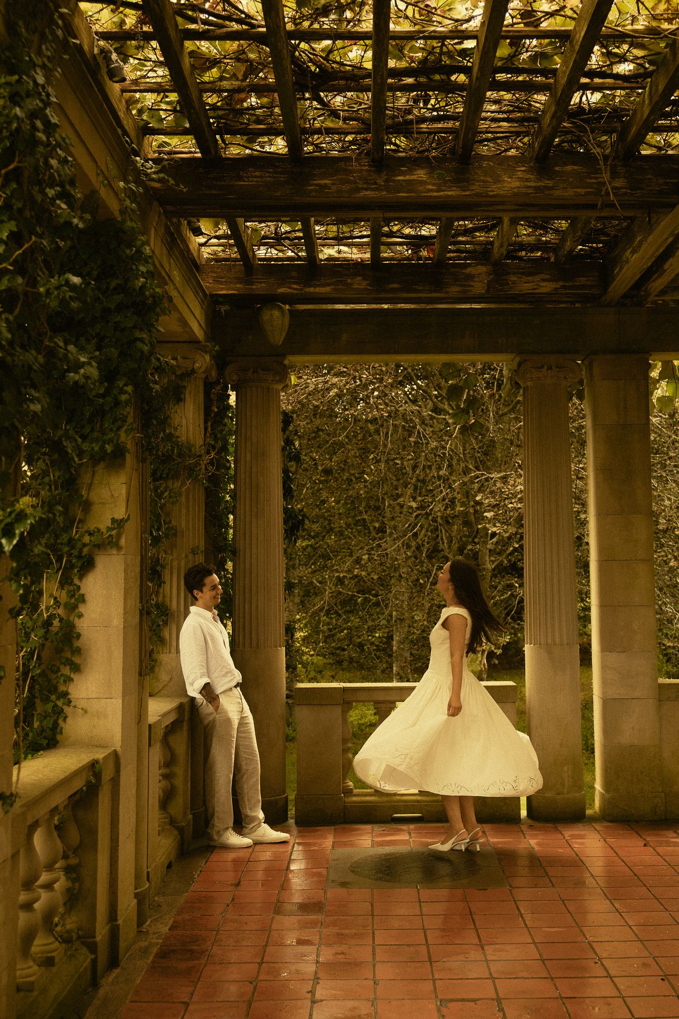 A couple in white attire stands apart under a vine-covered pergola with stone columns in a lush garden.